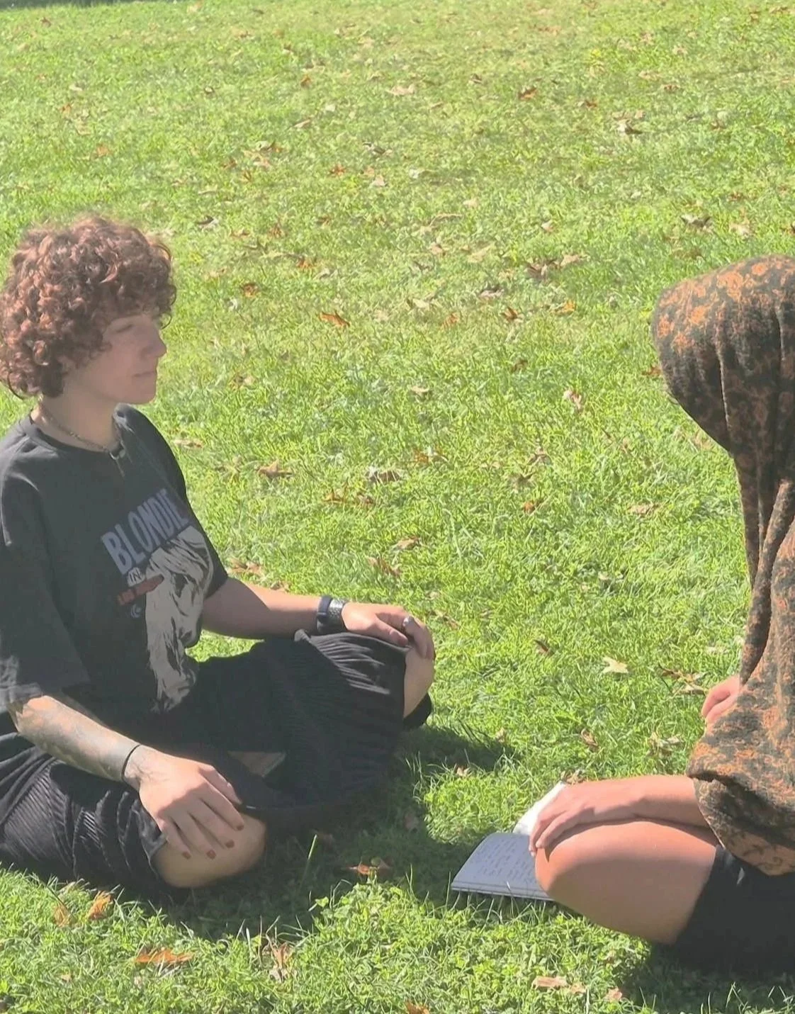 Two people sitting on grass outdoors with a notebook, one with curly hair and the other with a hooded jacket, engaged in conversation.