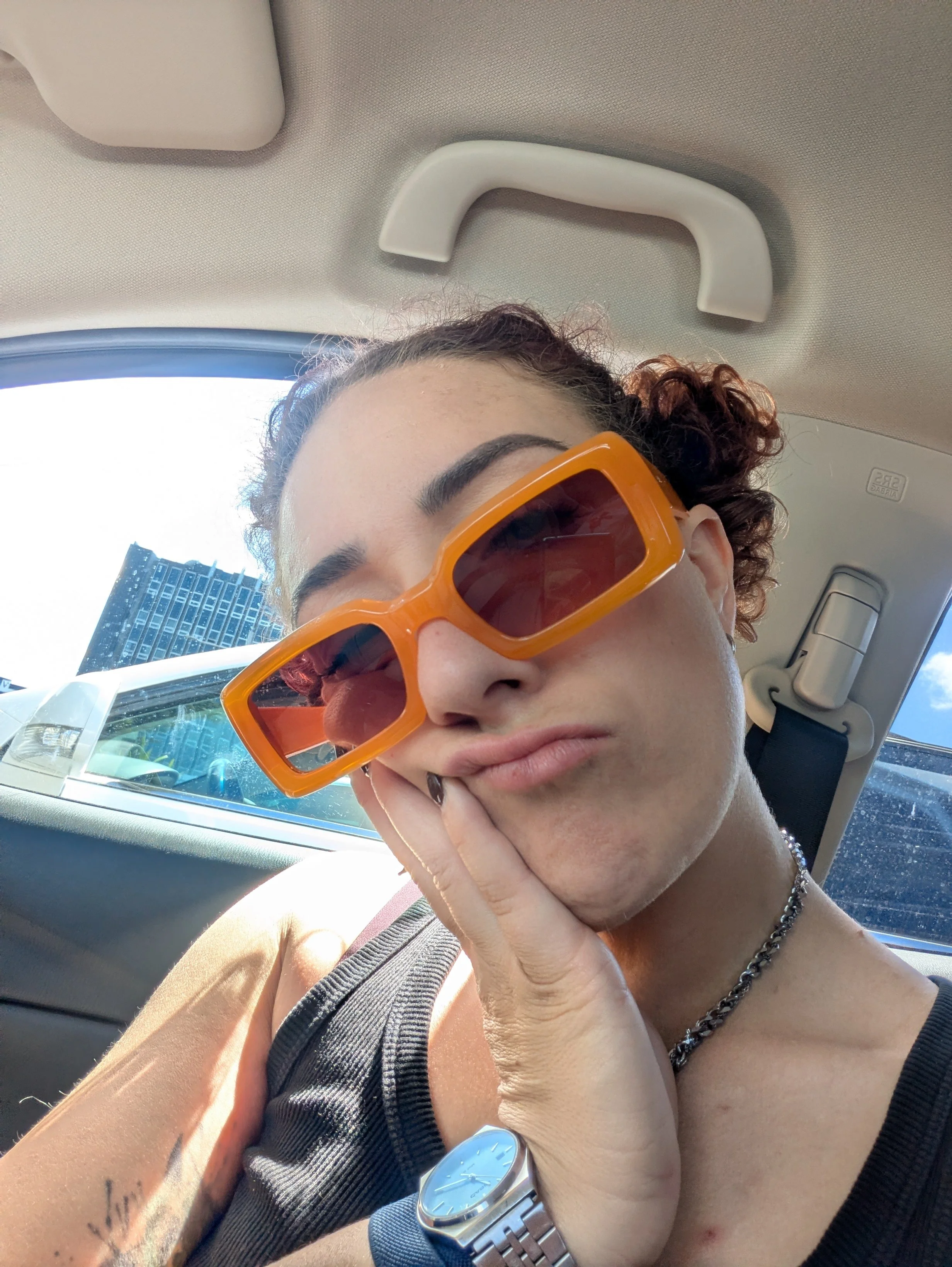A person with curly hair wearing large orange sunglasses, a watch, a chain necklace, and a striped tank top, sitting in a car with a cityscape visible through the window.