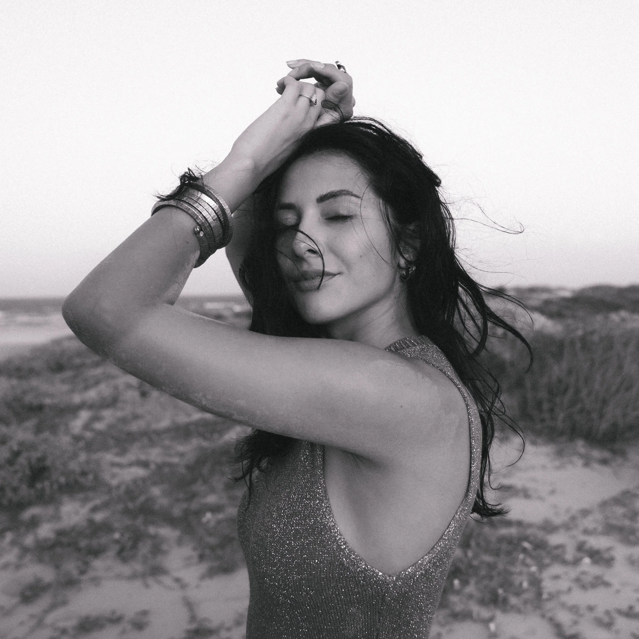 A black and white photograph of a woman with closed eyes holding her hand with rings above her head, wearing bangles, and a sleeveless, shimmering dress, in an outdoor setting.