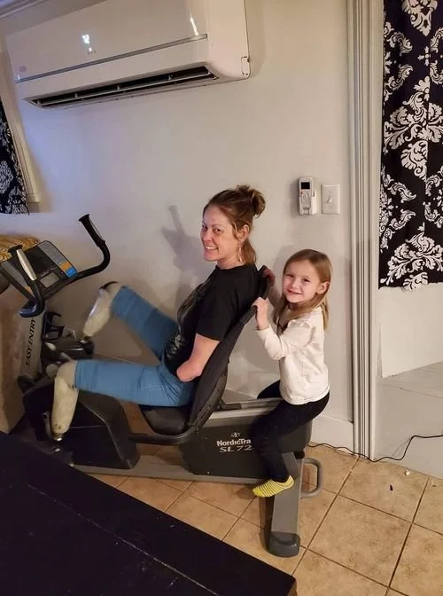 A woman sitting on a stationary exercise bike with a young girl standing behind her, holding onto her shoulders, both smiling. The woman is wearing black top and blue jeans, the girl is in a white top and black leggings, in a room with an air conditioner and patterned curtains.
