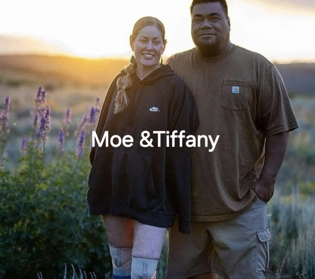 A woman and a man standing outdoors during sunset, with purple flowers in the background. The woman is smiling and wearing a black Nike hoodie, and the man is wearing a brown Carhartt T-shirt. Text over the image reads 'Moe & Tiffany.'