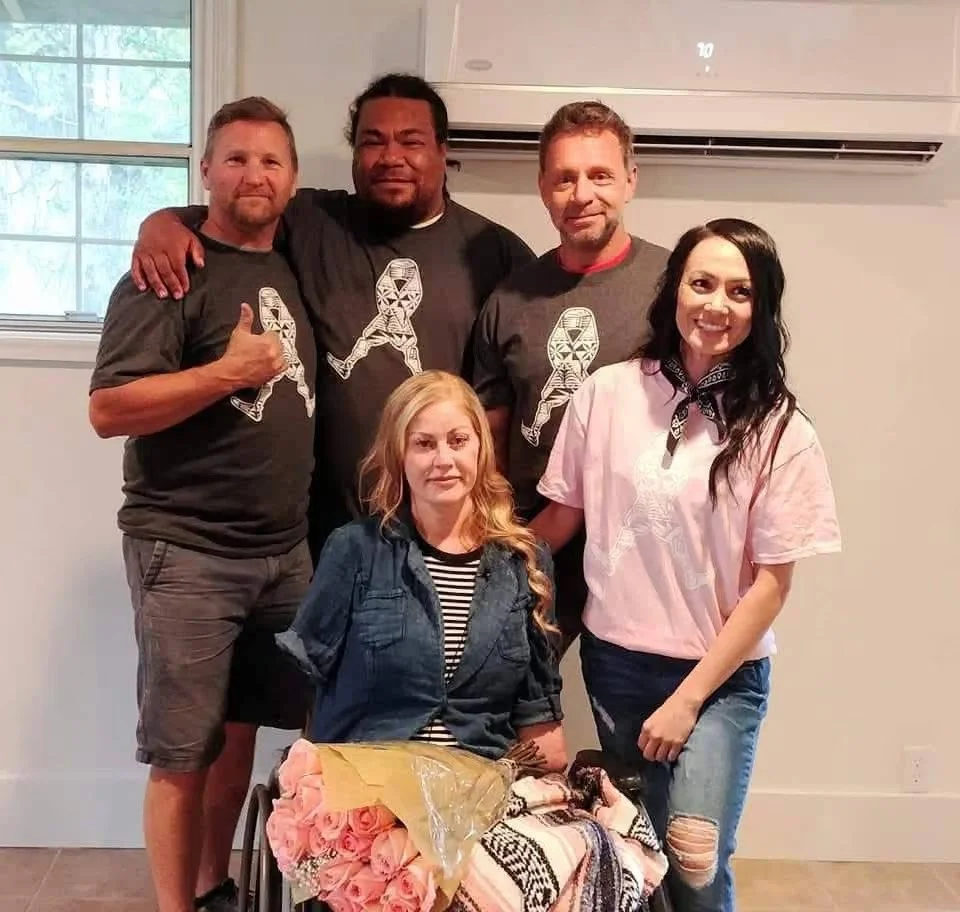 A group of five people indoors, one woman in a wheelchair with a bouquet of pink roses, with four people standing behind her, all smiling and posing for a photo.