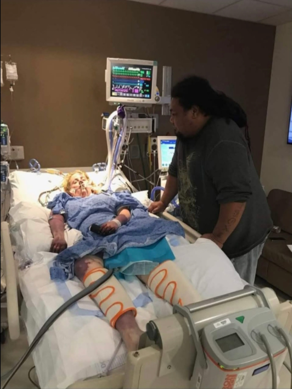 A woman in a hospital bed connected to medical devices, with a person beside her holding her hand.