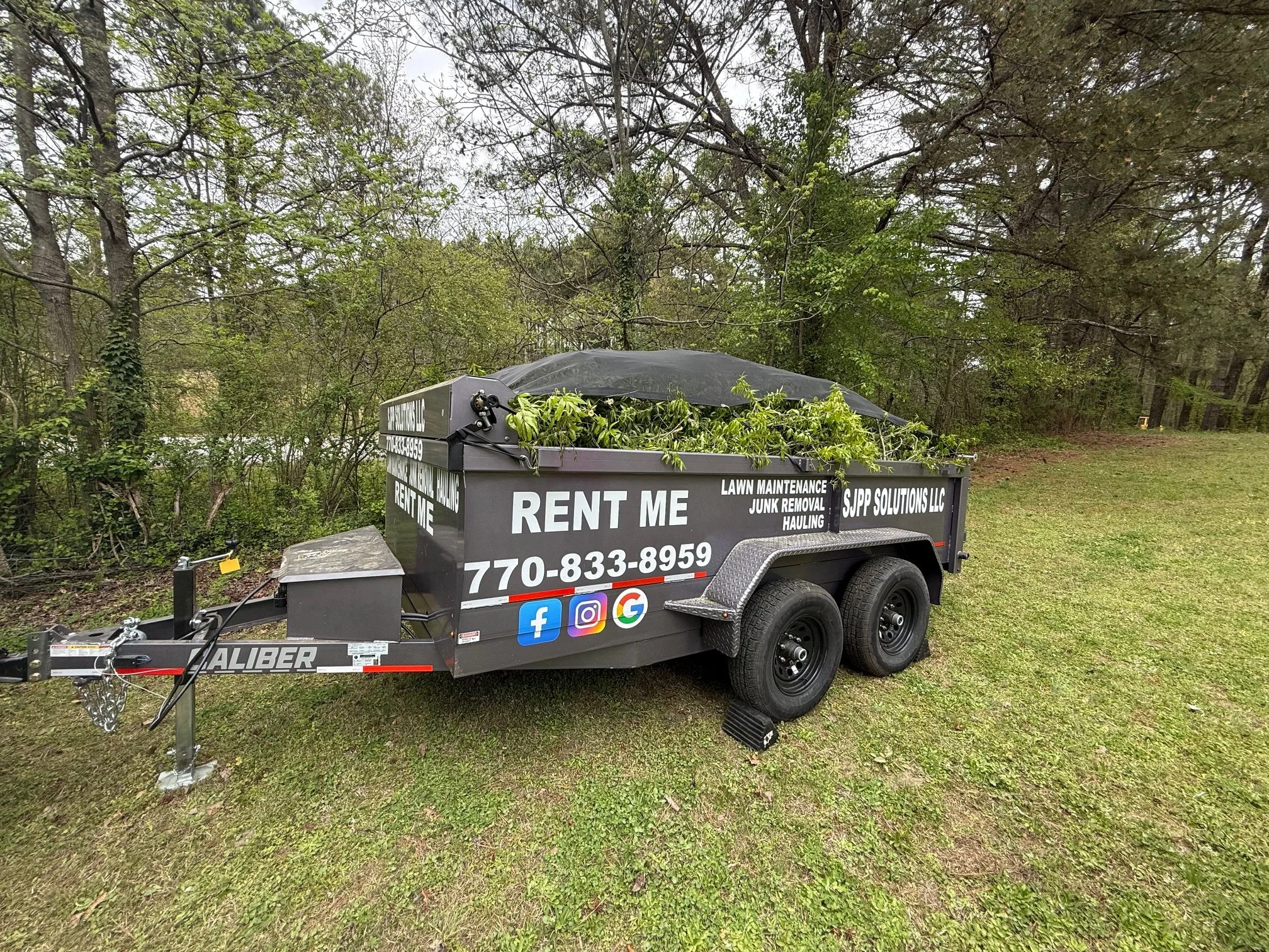 A black utility trailer filled with fresh green tree and shrub trimmings parked on grass in a wooded area.