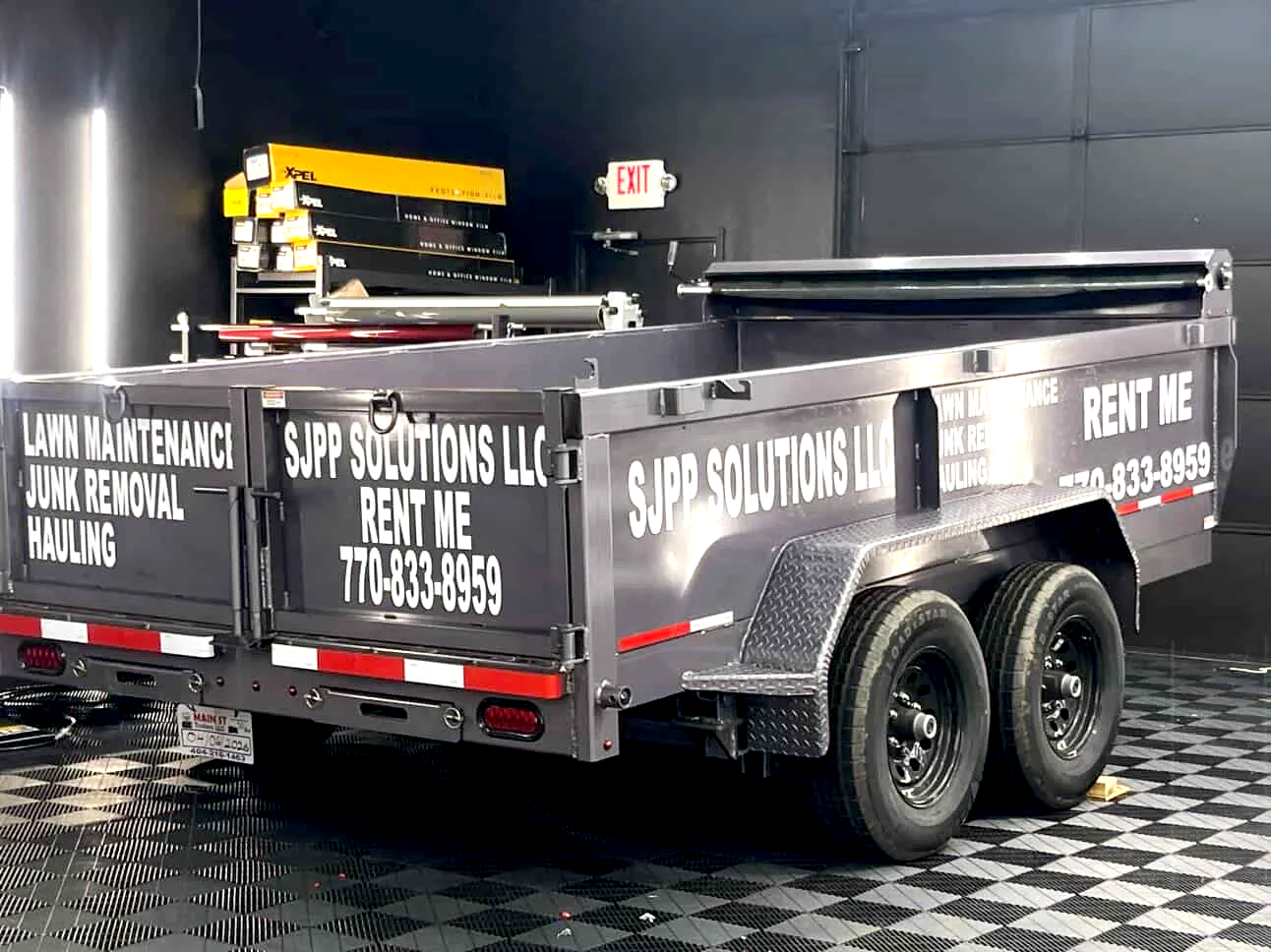 A double-axle utility trailer with black and white lettering advertising SJP Solutions LLC for lawn maintenance, junk removal, and hauling services, parked on a checkered black and white floor inside a garage or warehouse.