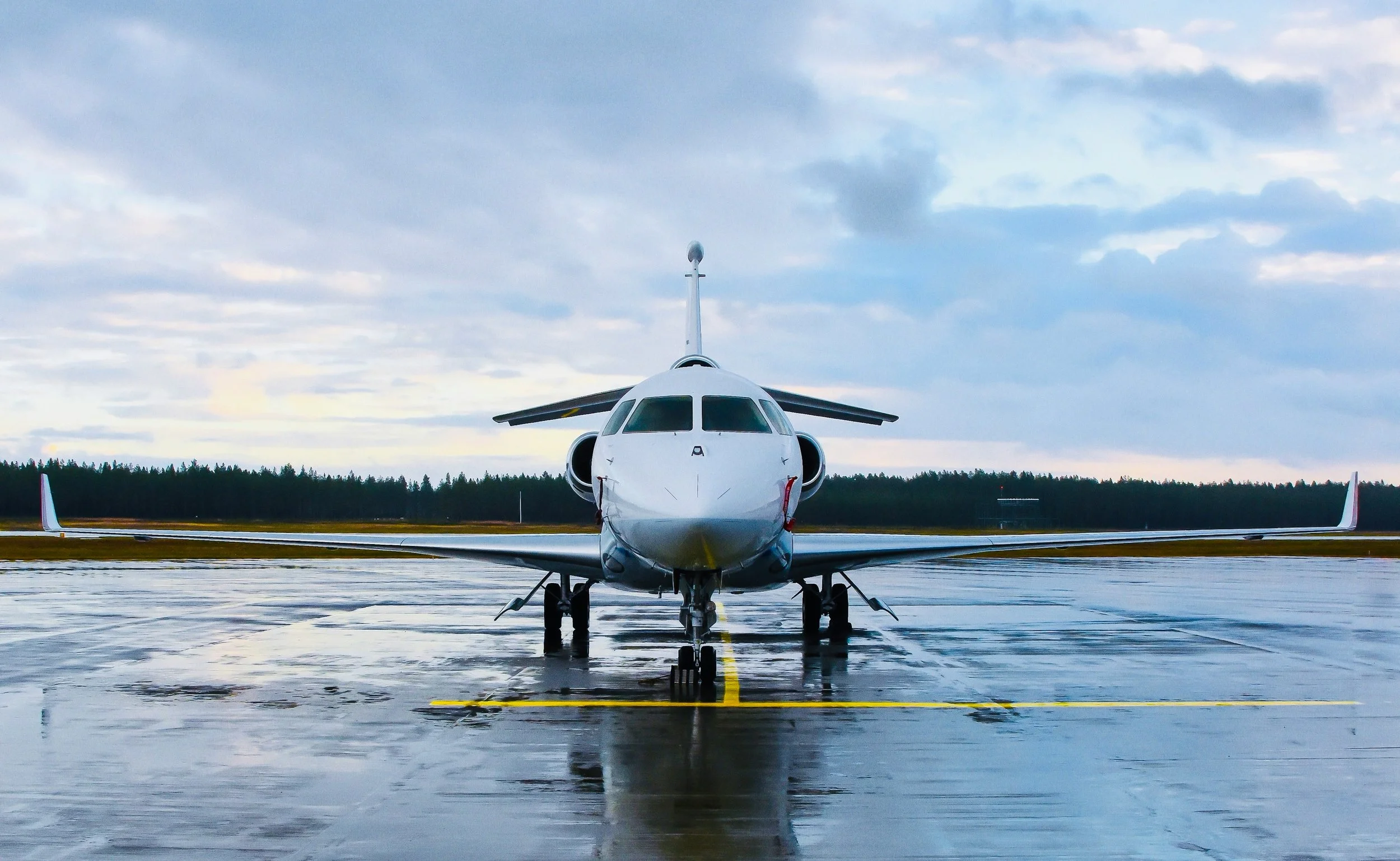 A white airplane is parked on a wet runway under a cloudy sky, with trees in the background.