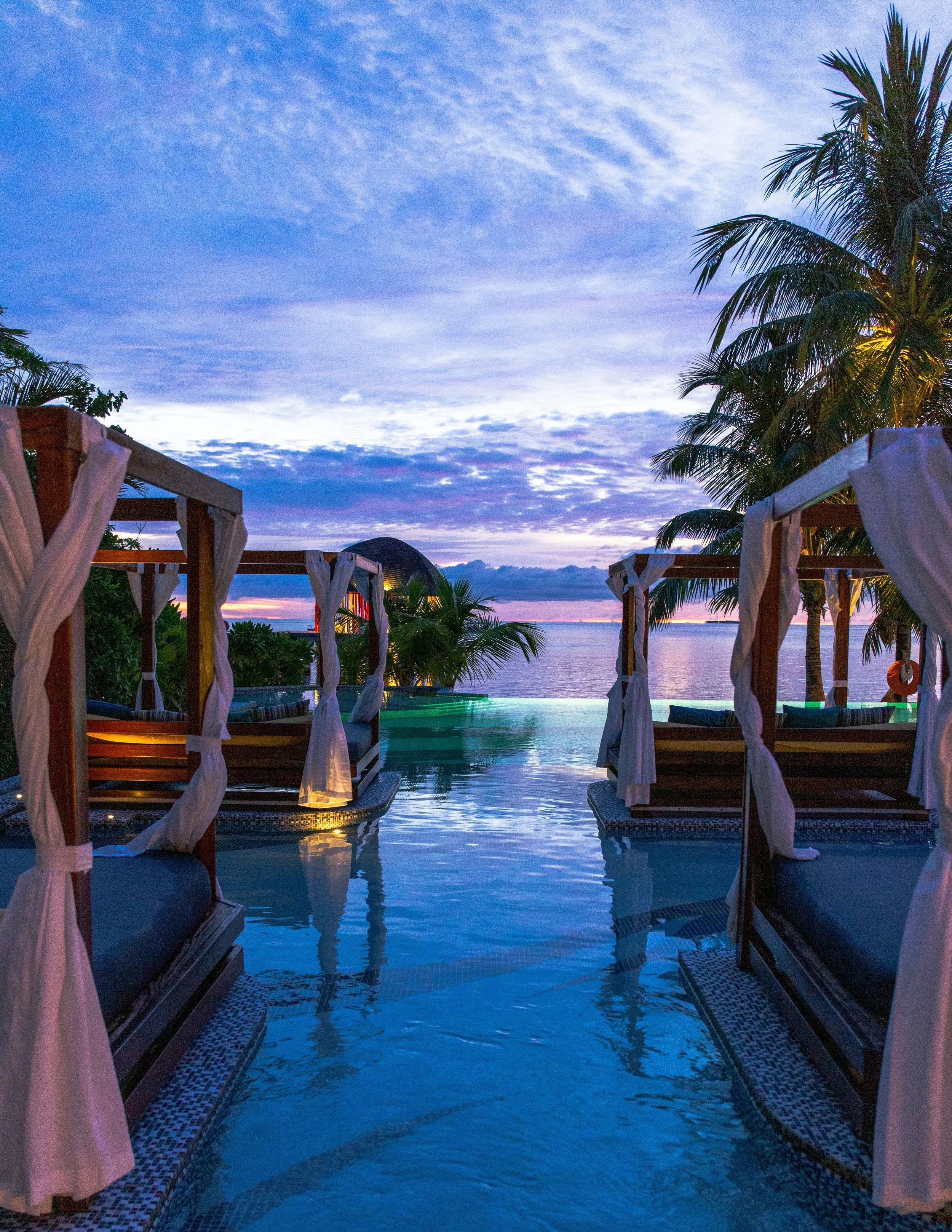 Poolside cabanas with white curtains overlooking a calm body of water during dusk, with palm trees and a colorful evening sky.