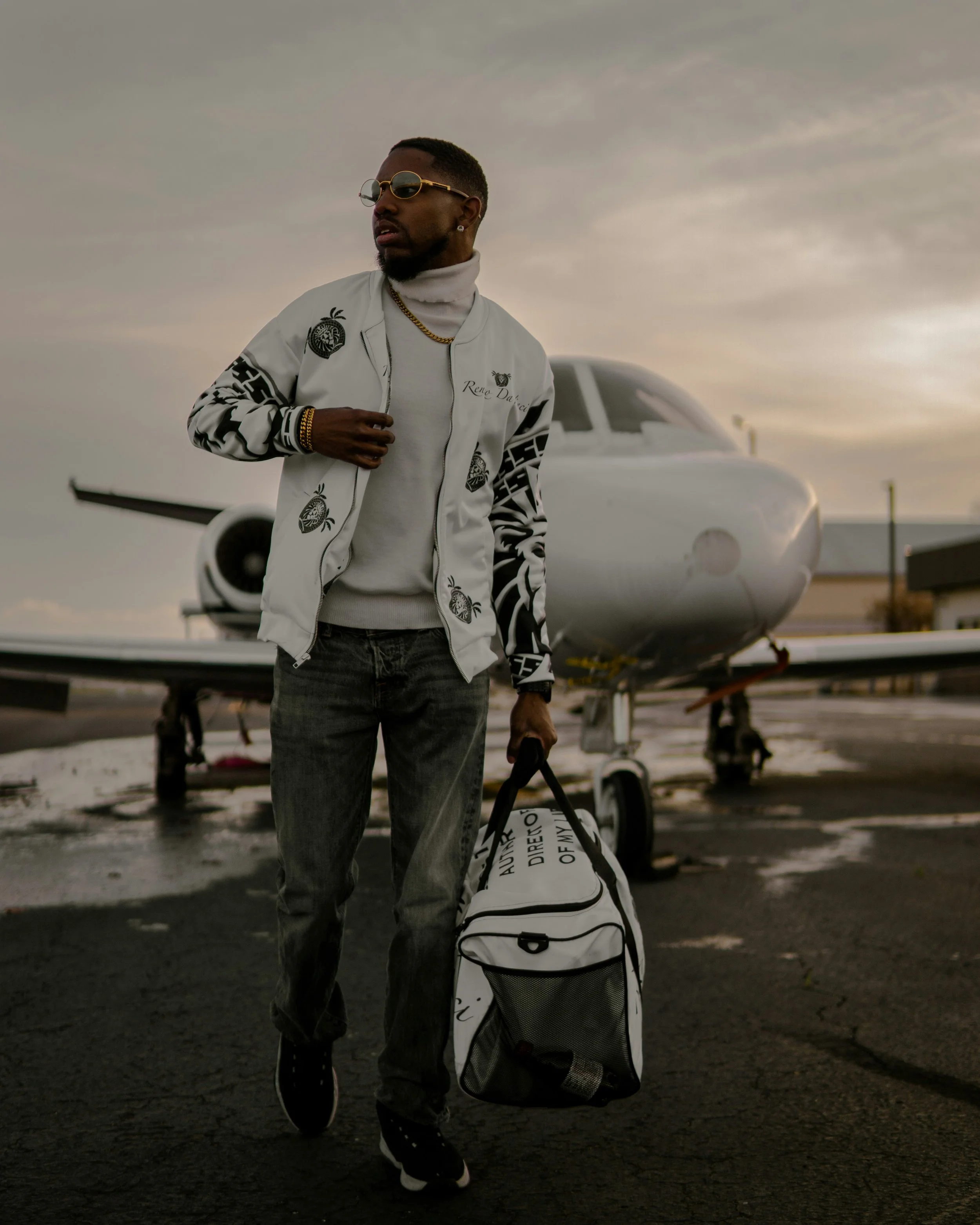 A man walking beside a white private jet at an airport with a cloudy sky, carrying a white golf bag and dressed in casual sportswear, including sunglasses, a white jacket with black patterns, a chain, and a turtleneck shirt.
