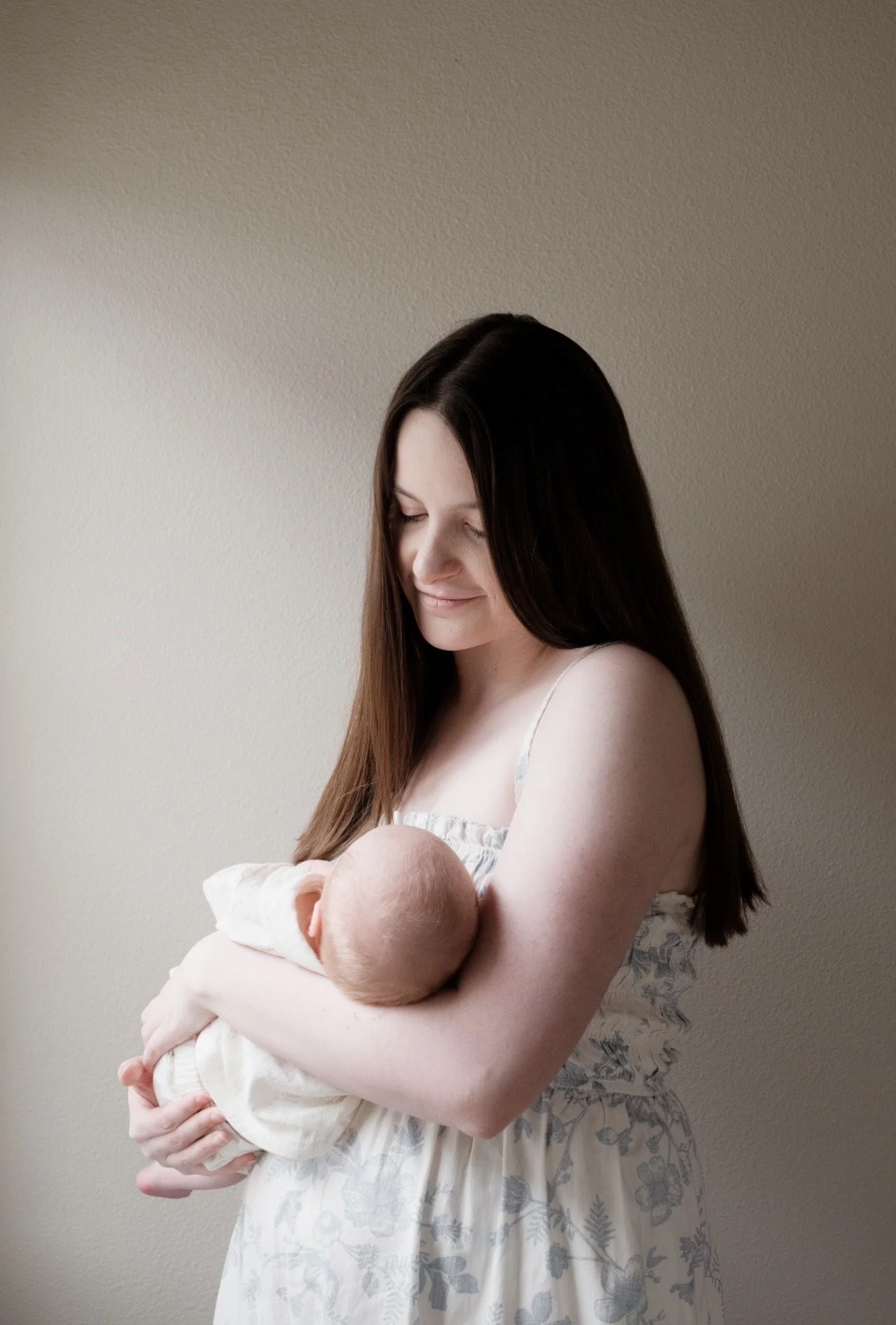 A woman with long dark hair holding a newborn baby wrapped in a blanket, standing against a plain wall, smiling gently with her eyes closed.