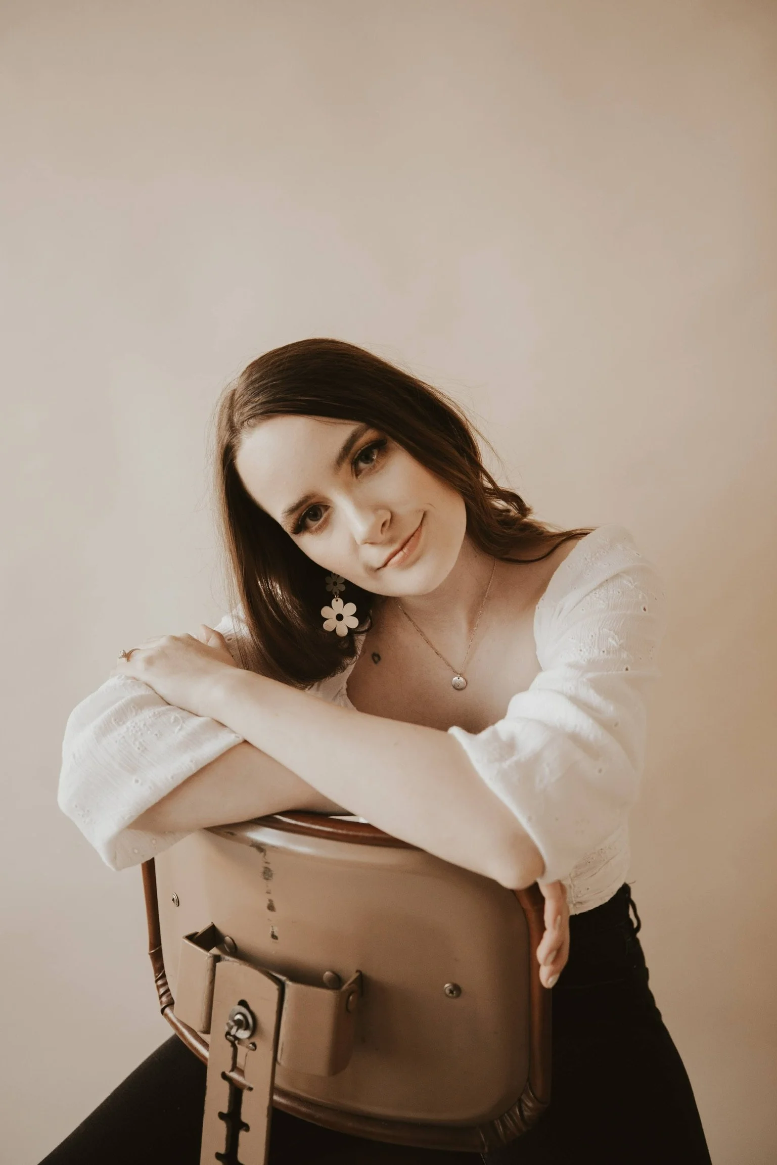 A young woman with brown hair and fair skin sitting on a chair, smiling at the camera, wearing a white top and floral earrings.