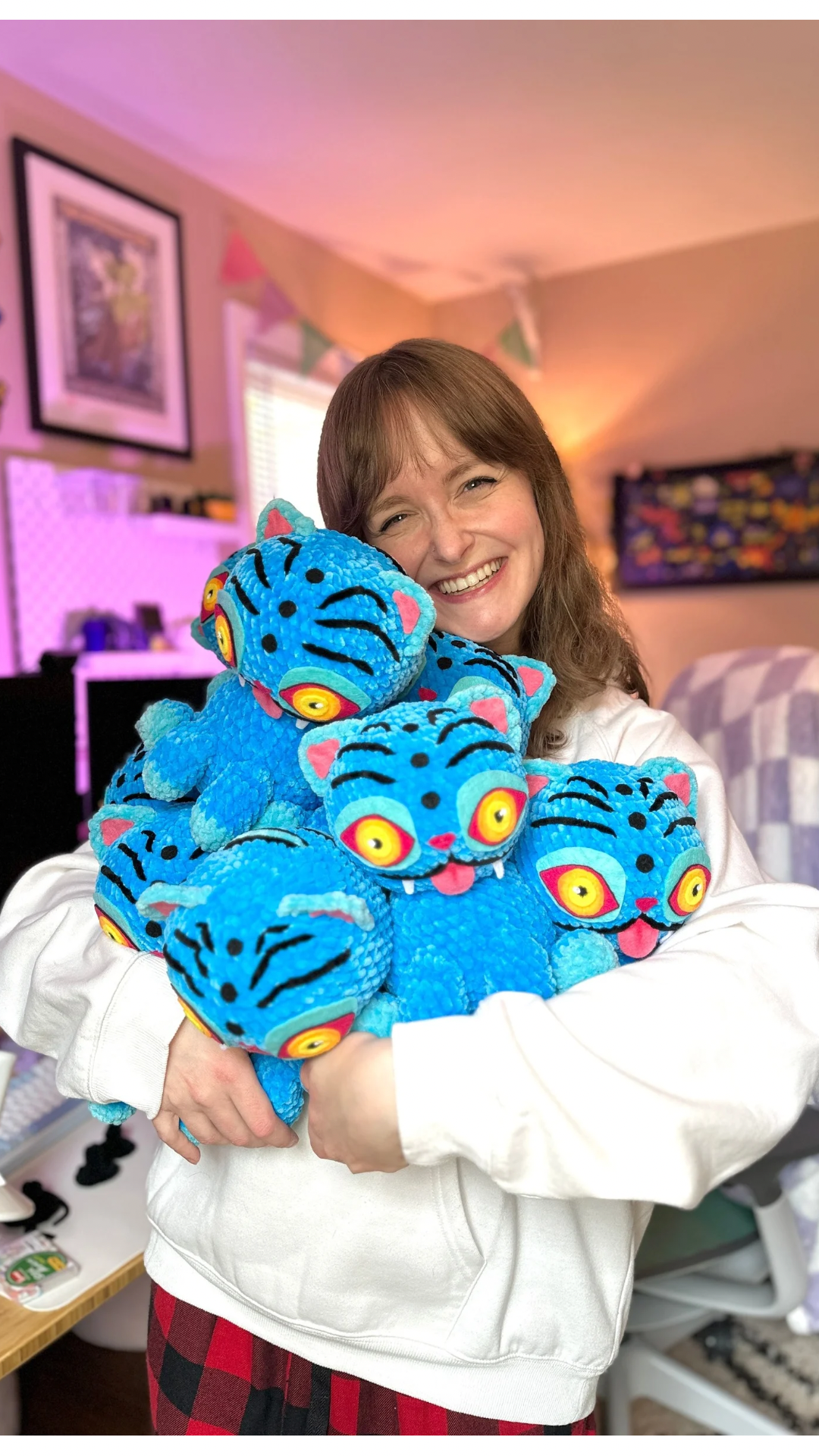 A woman with brown hair holding multiple plush toys resembling blue monsters with big yellow eyes and pink accents, smiling in a colorful, decorated room.
