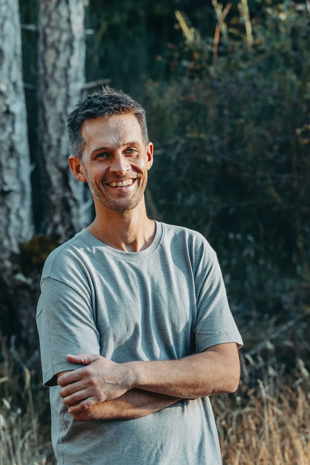 A smiling man with short, dark hair and a light beard, standing outdoors in a forested area. He is wearing a light gray t-shirt and has his arms crossed. The background features trees and natural foliage.