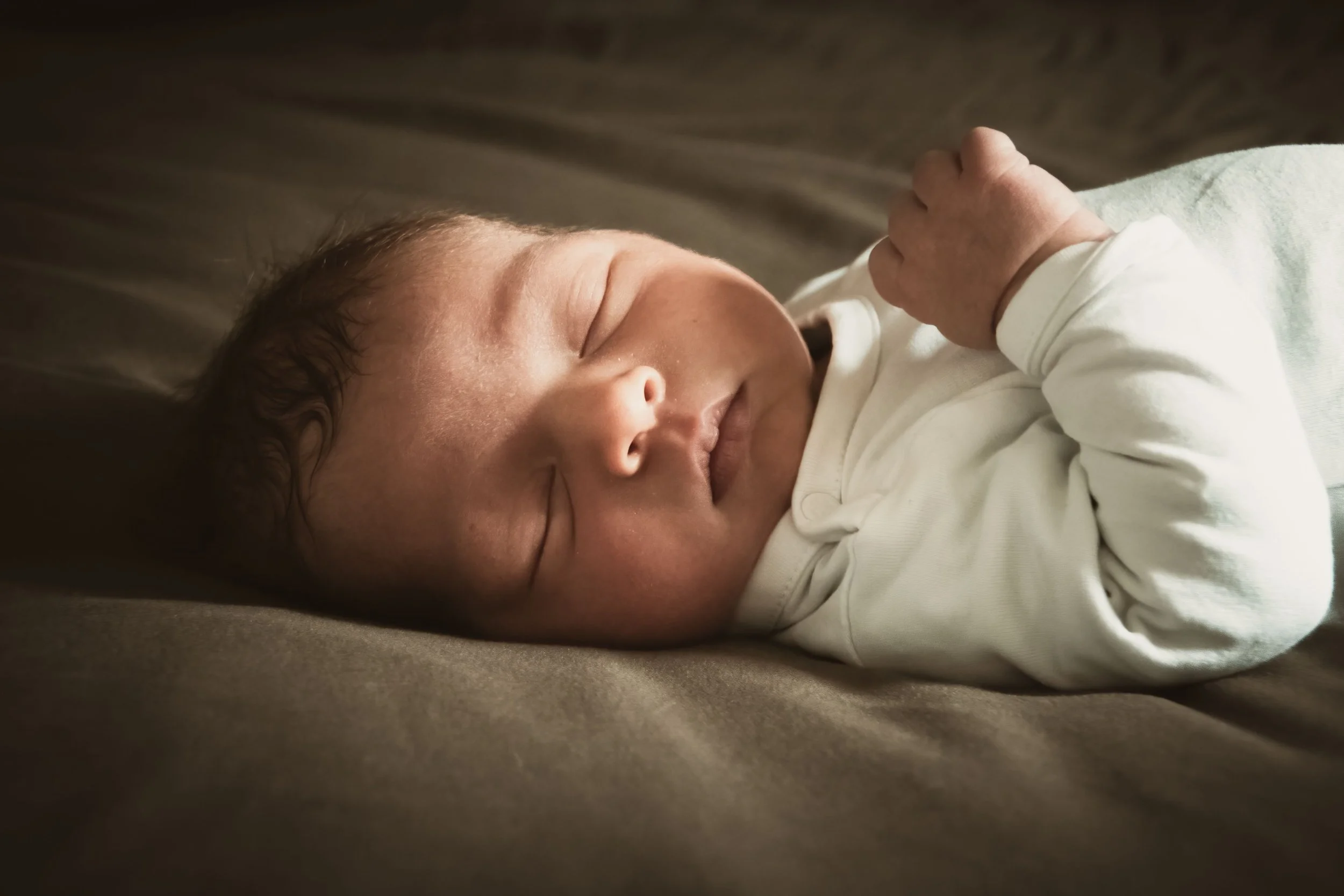 Close-up of a newborn baby peacefully sleeping on a soft surface, dressed in a white long-sleeve outfit, with a gentle expression and hands relaxed.