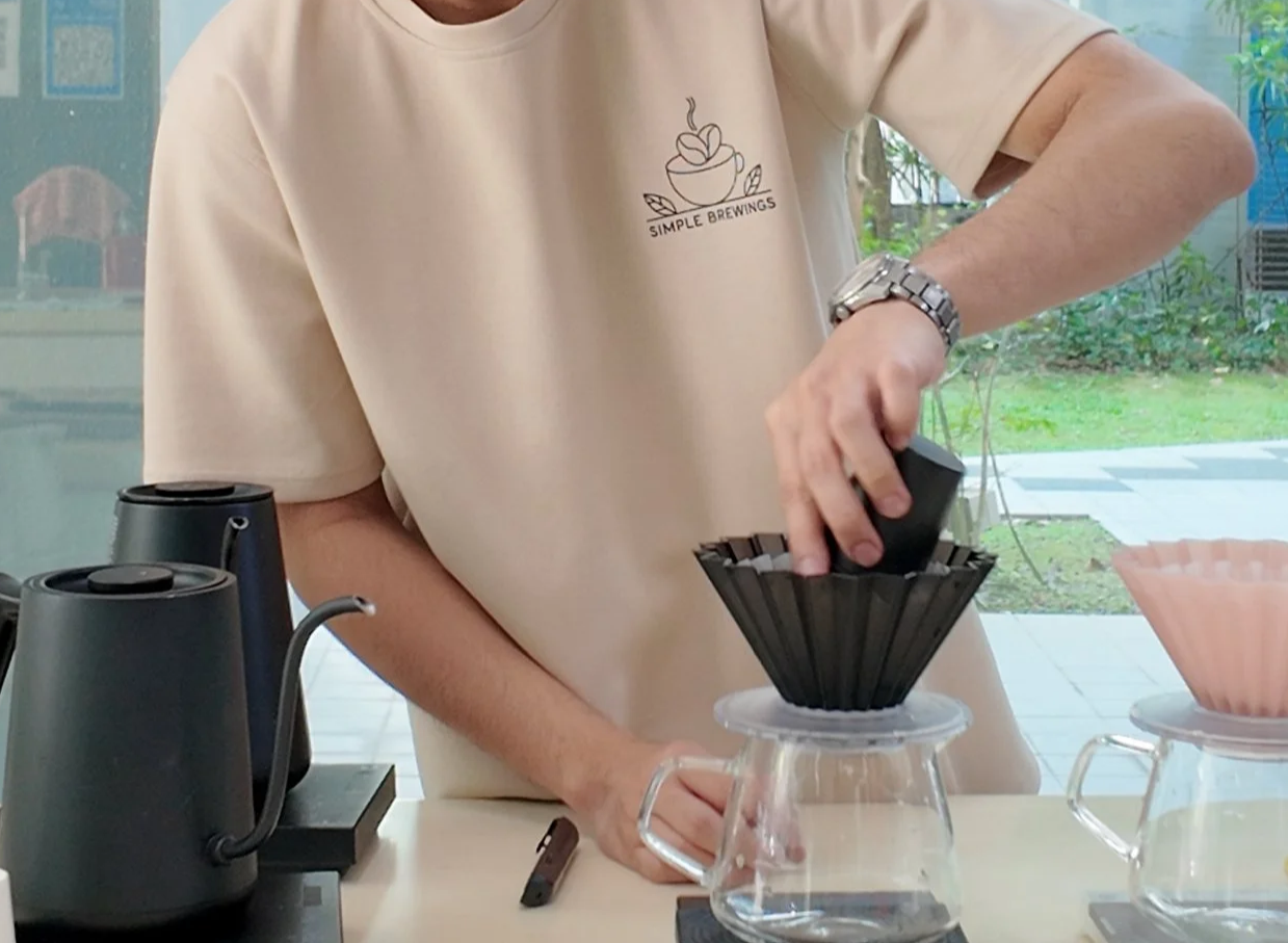 Person pouring freshly brewed coffee through a black coffee dripper into a glass carafe, with coffee-making equipment and materials on the table.