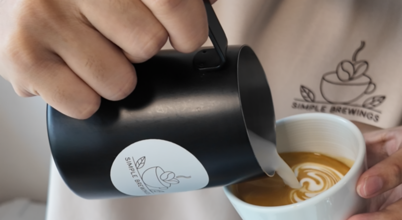 A person pouring milk into a cup of coffee with latte art, wearing a shirt with a coffee logo.