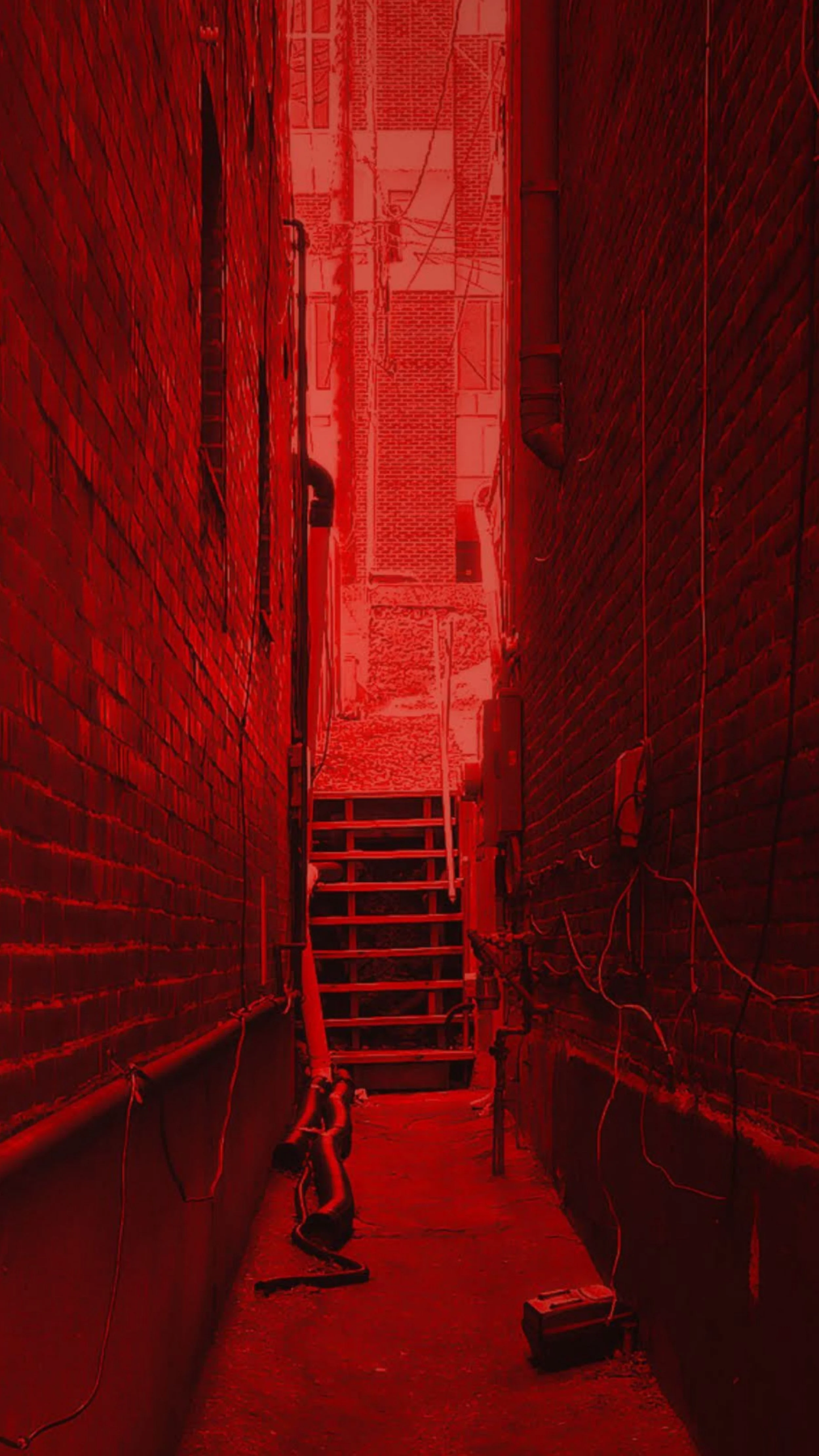Narrow alleyway between two brick buildings, with various wires and cables along the walls, a ladder in the middle, and a toolbox on the ground, all illuminated in red light.