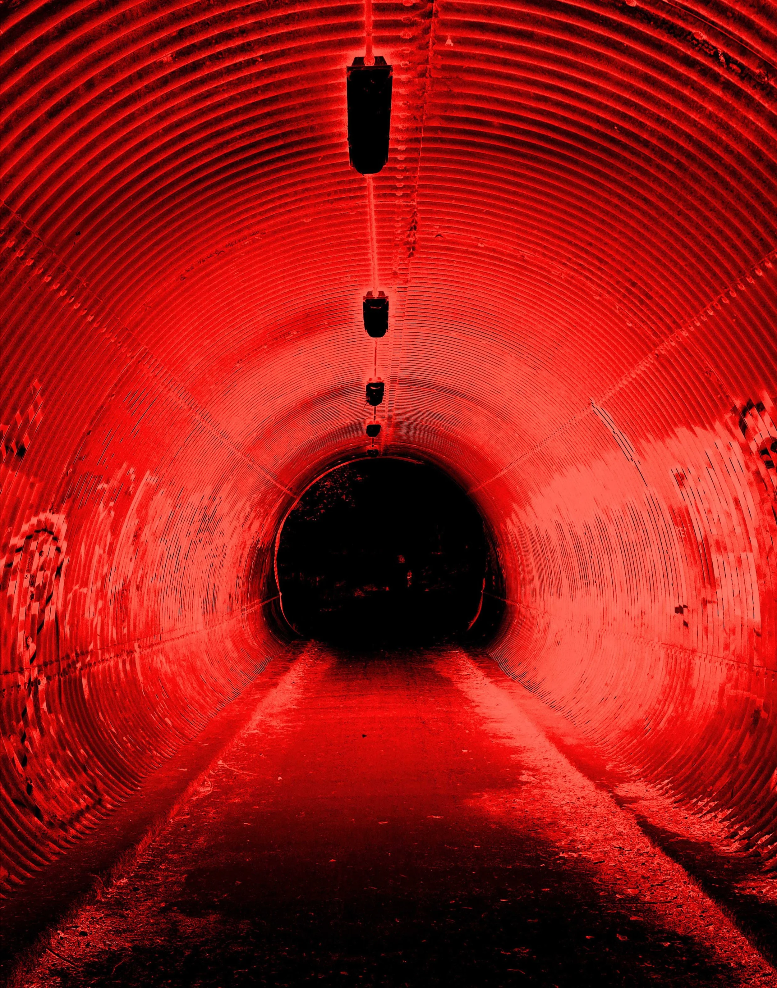 A dark tunnel illuminated with red lighting, with evenly spaced black lights hanging from the ceiling, converging towards a dark opening at the end.