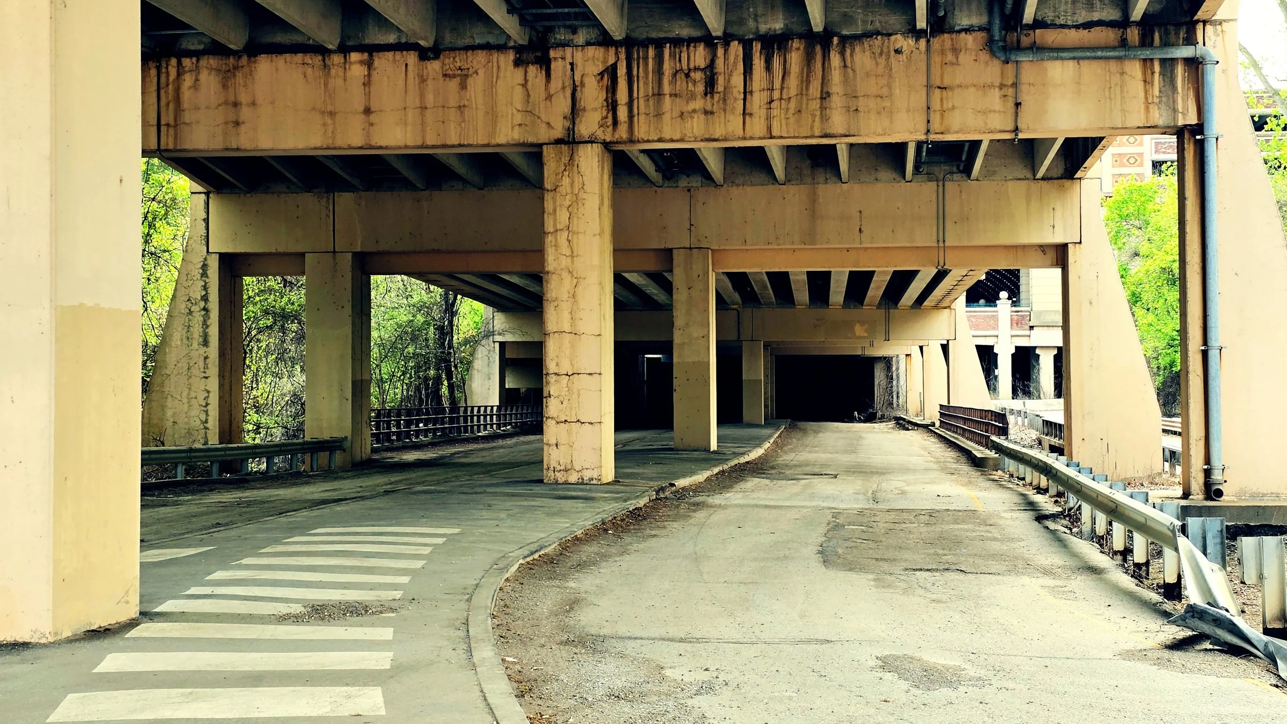 Underpass with concrete pillars and a road curving underneath, surrounded by trees.