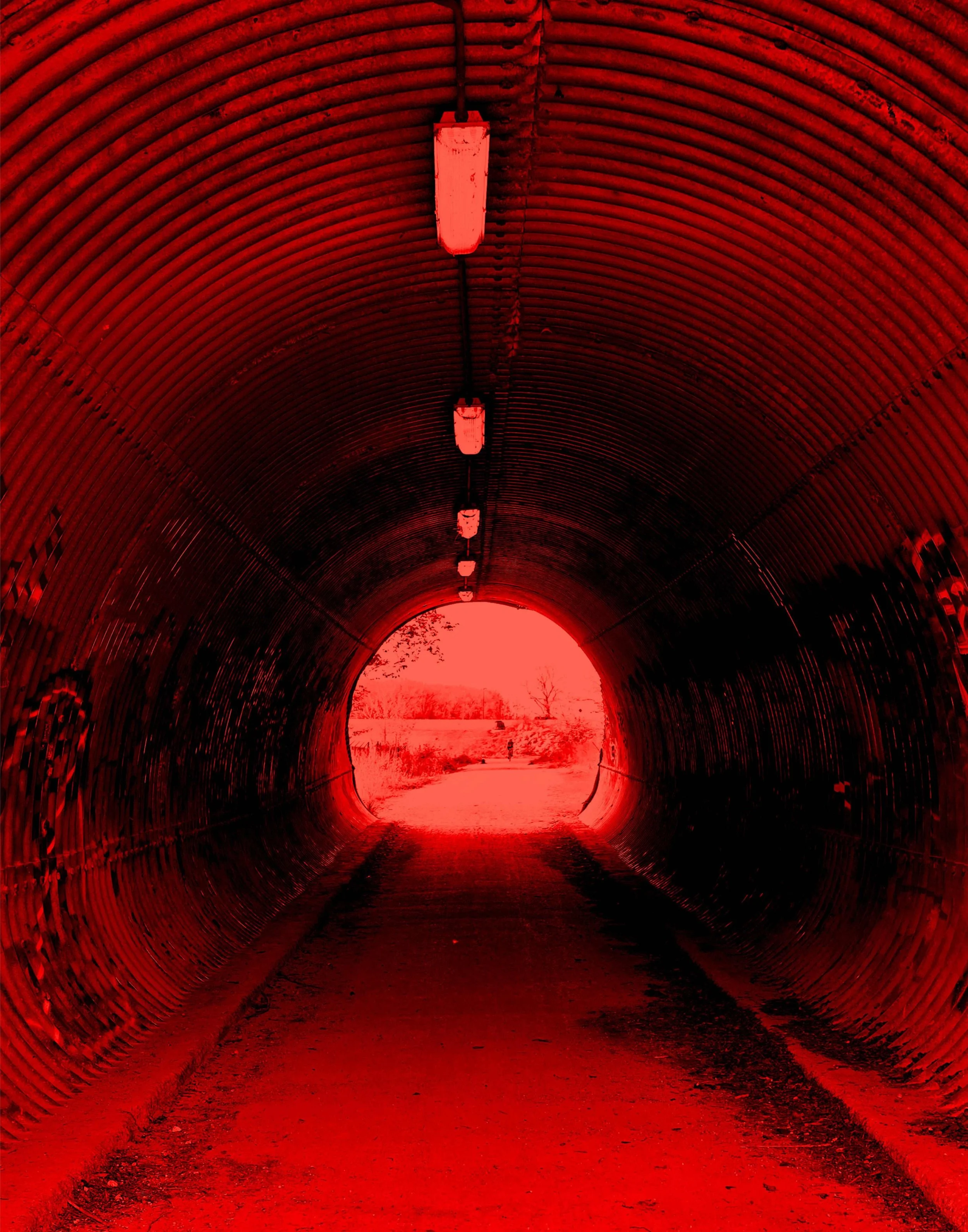 Long tunnel with metal ribs painted red, illuminated by fluorescent lights, leading to an open exit with a view of a rural landscape with trees and a field.