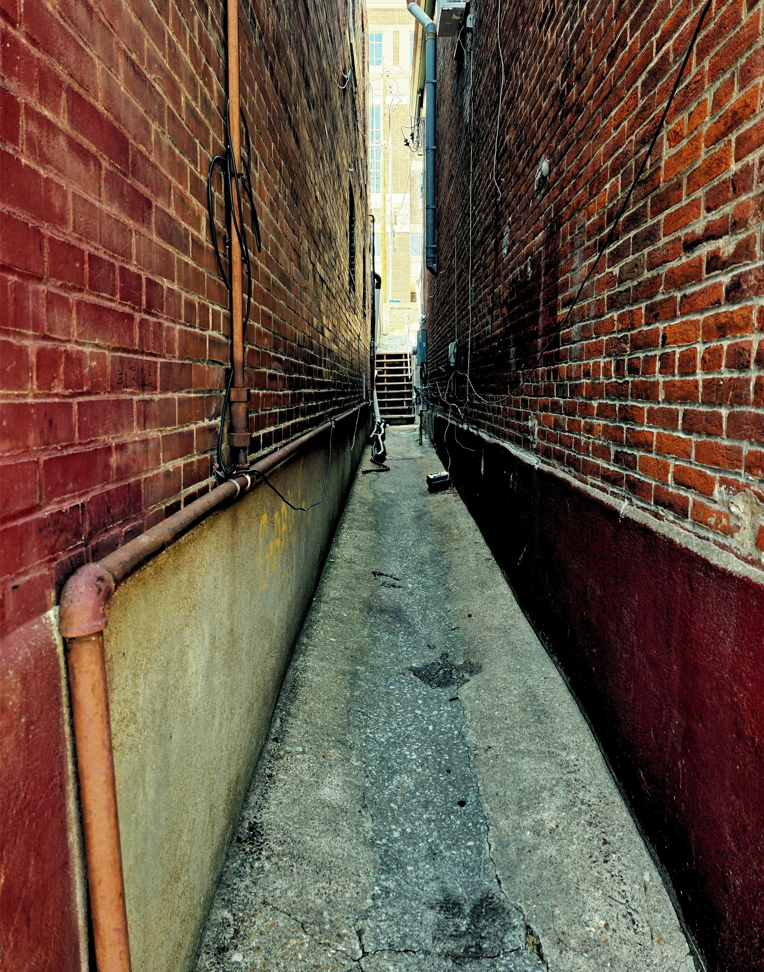 A narrow alleyway between two brick buildings with exposed pipes and wires, leading to stairs and an open area in the distance.