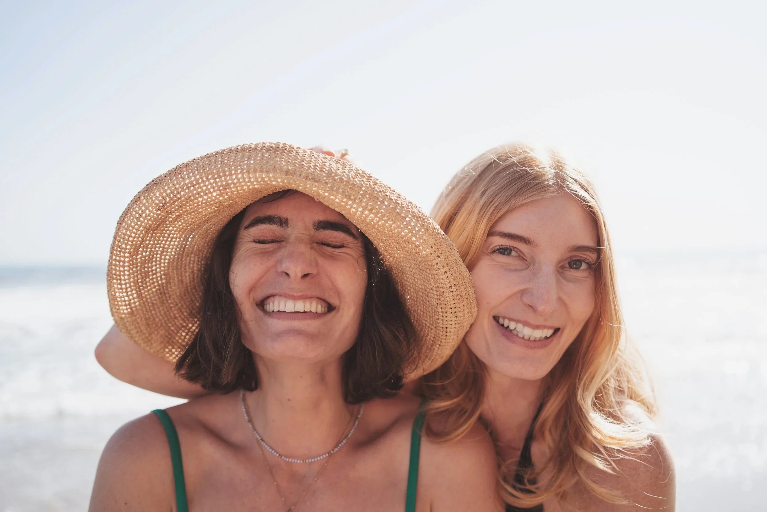 Two women at the beach, one wearing a large sunhat and smiling with eyes closed, the other with red hair, smiling at the camera, with ocean in the background.