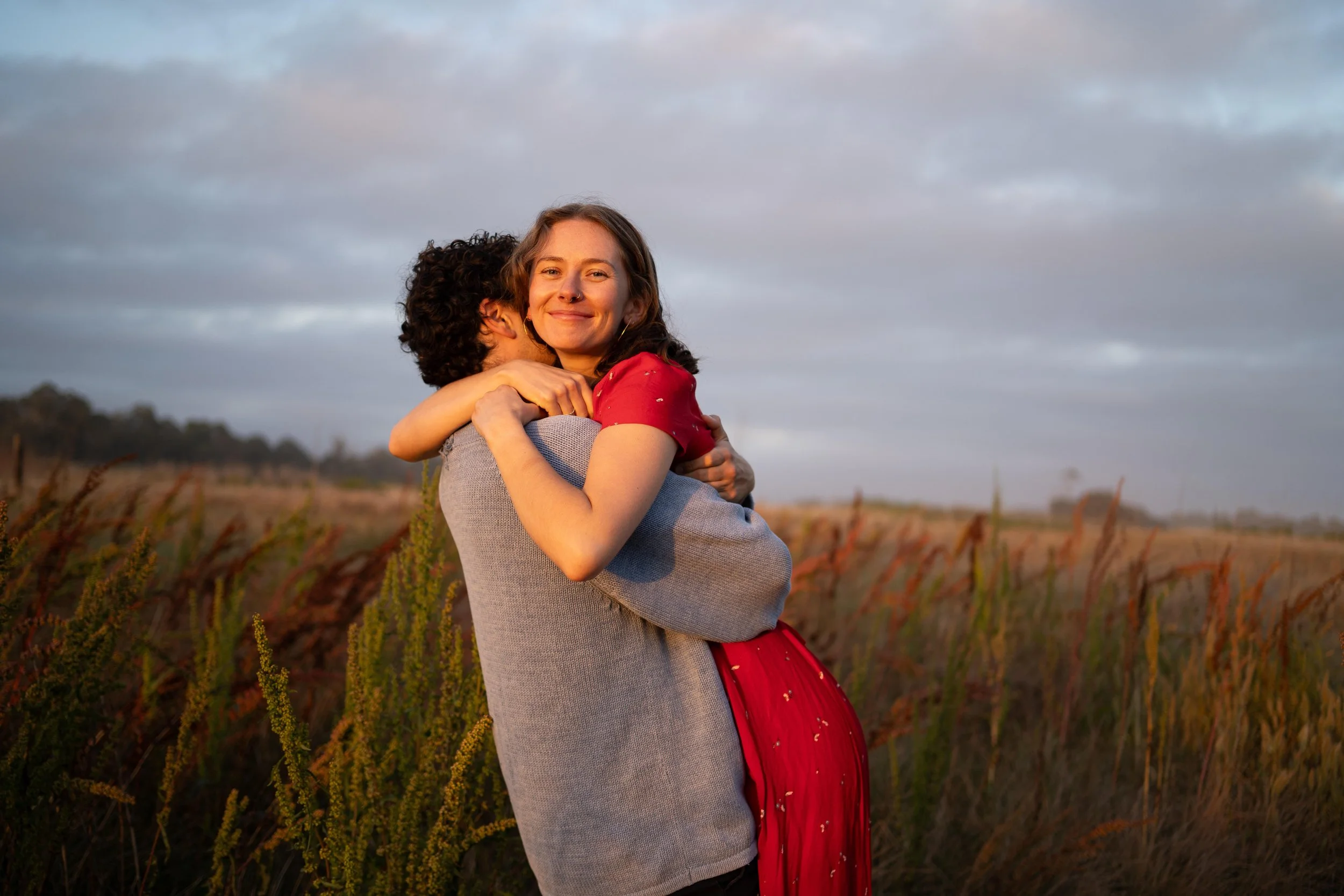 Two women hugging in a field at sunset, one woman smiling at the camera while the other has her face turned away.