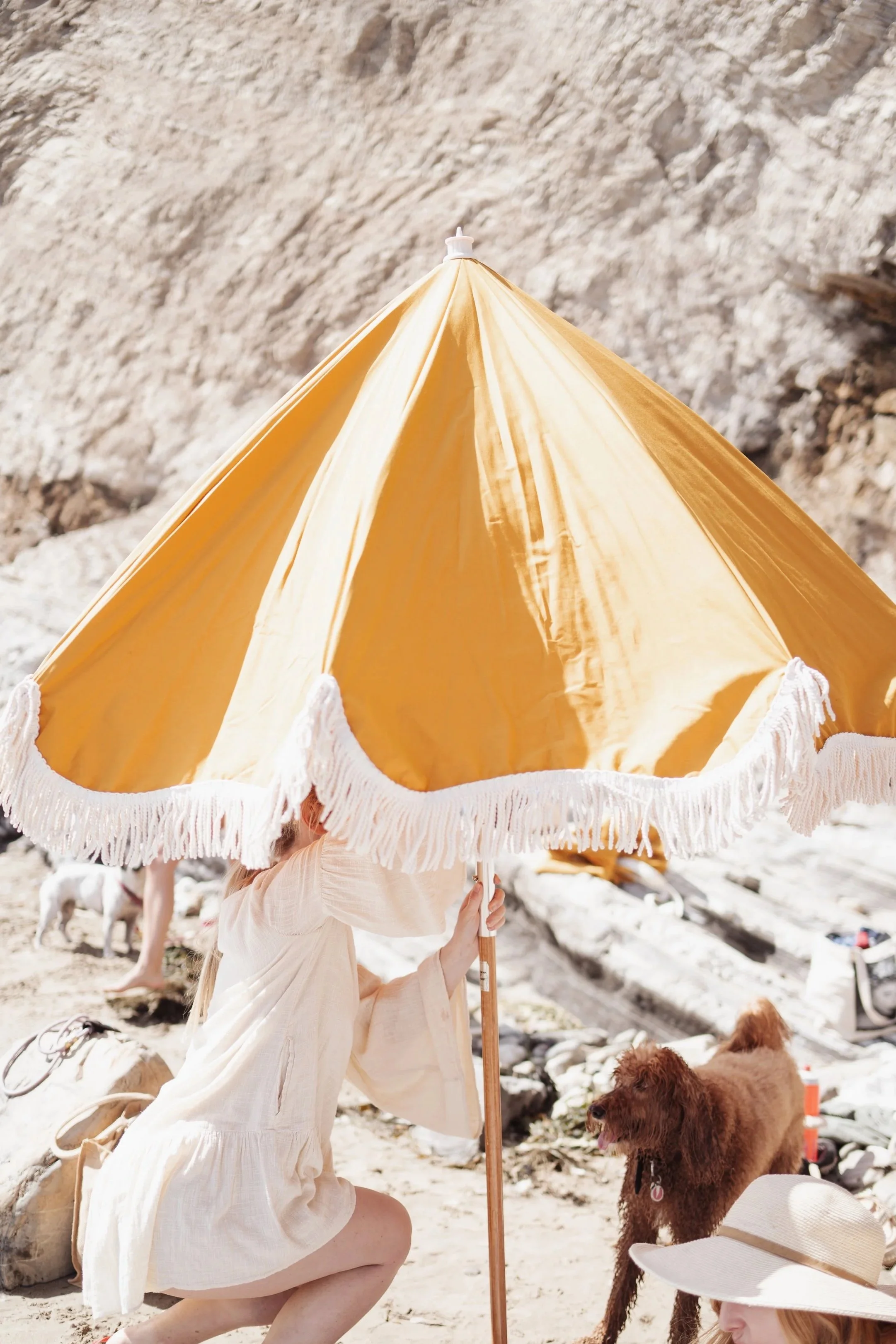 A woman kneeling on a beach under a yellow umbrella with a white fringe, holding the umbrella pole. There are brown dogs near her, one sitting and another close by. The background shows a rocky cliff and beach scenery.