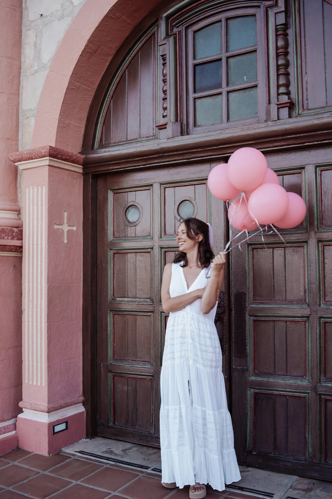 Bachelorette party Santa Barbara. Woman smiling and holding pink balloons in front of a large wooden arched door with decorative woodwork and stained glass panels, on a pink stucco building with a cross symbol and a tiled sidewalk.