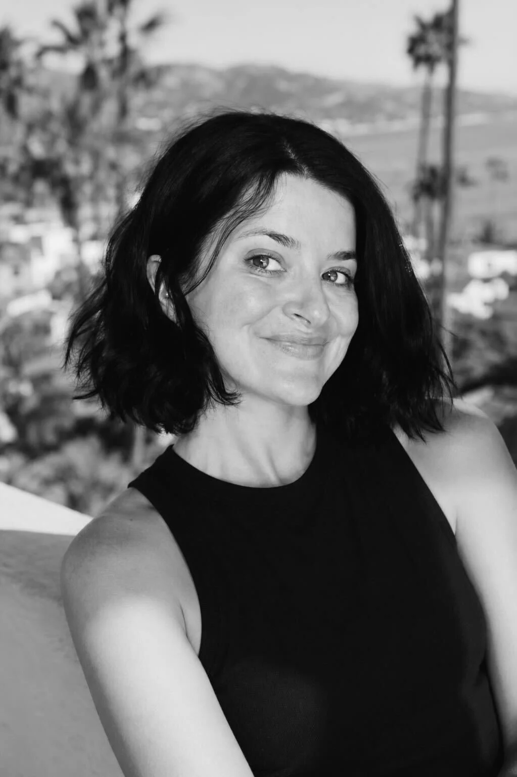Black and white photo of a woman with short dark hair, smiling, wearing a sleeveless black top, with a background of palm trees and distant hills.