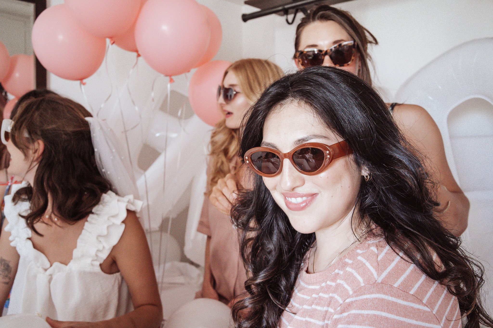 Group of women wearing sunglasses, smiling, and sitting around a table decorated with pink balloons at a celebration or party.