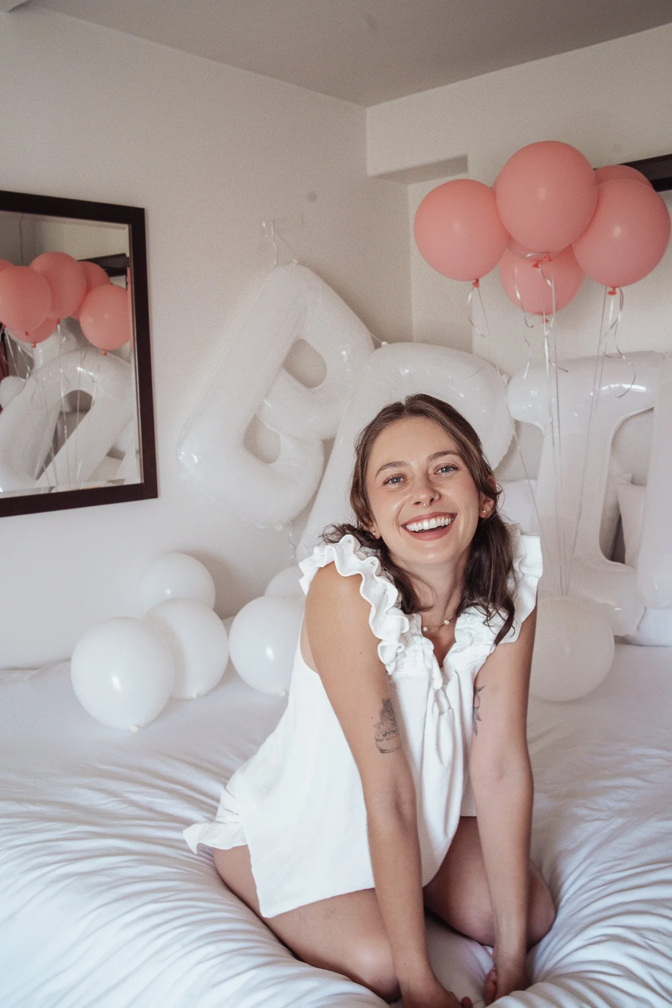 A young woman with brown hair, smiling, sitting on a bed surrounded by white balloons and pink balloons. A mirror and a wall with white paint are visible in the background.