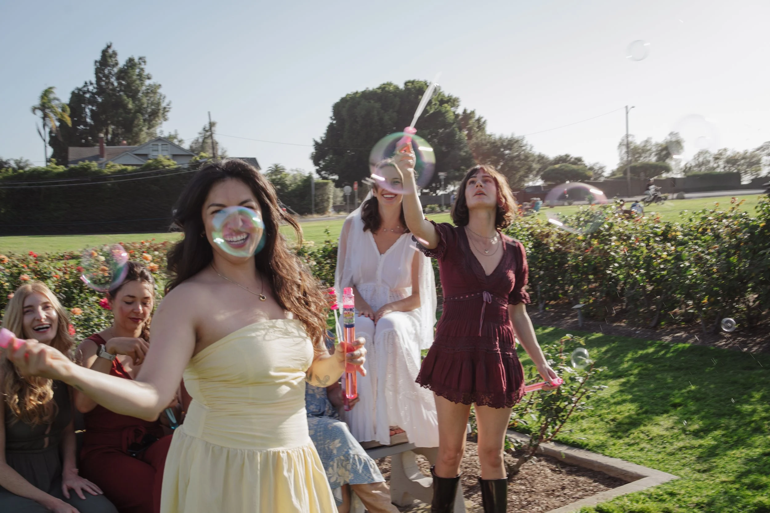 A group of women outdoors, with some holding bubble wands, smiling and enjoying a sunny day in a park or garden.
