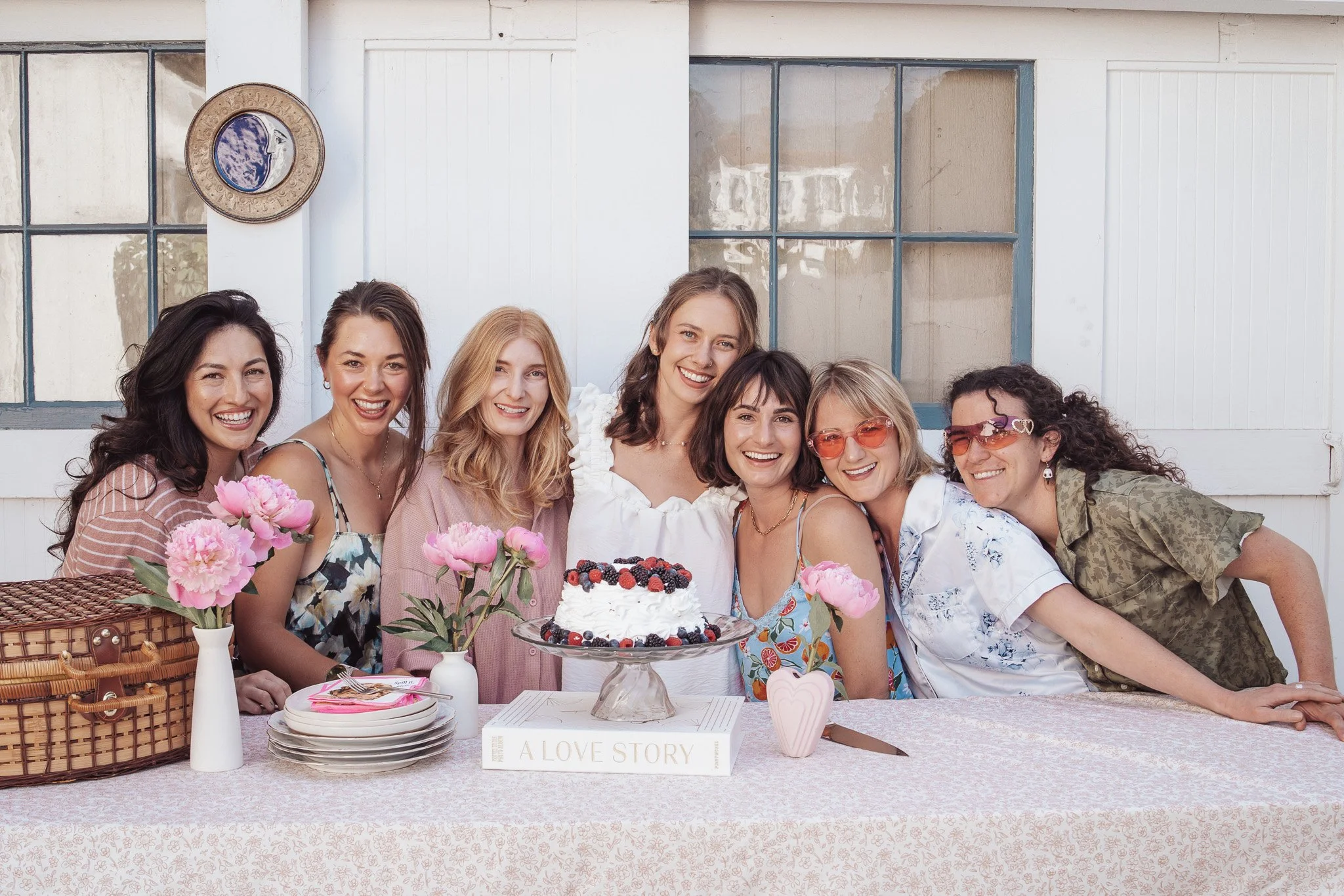 A group of eight women smiling at a birthday celebration with a cake decorated with berries, pink flowers, and a pink heart-shaped decoration. They are gathered around a table outdoors with a white building in the background.