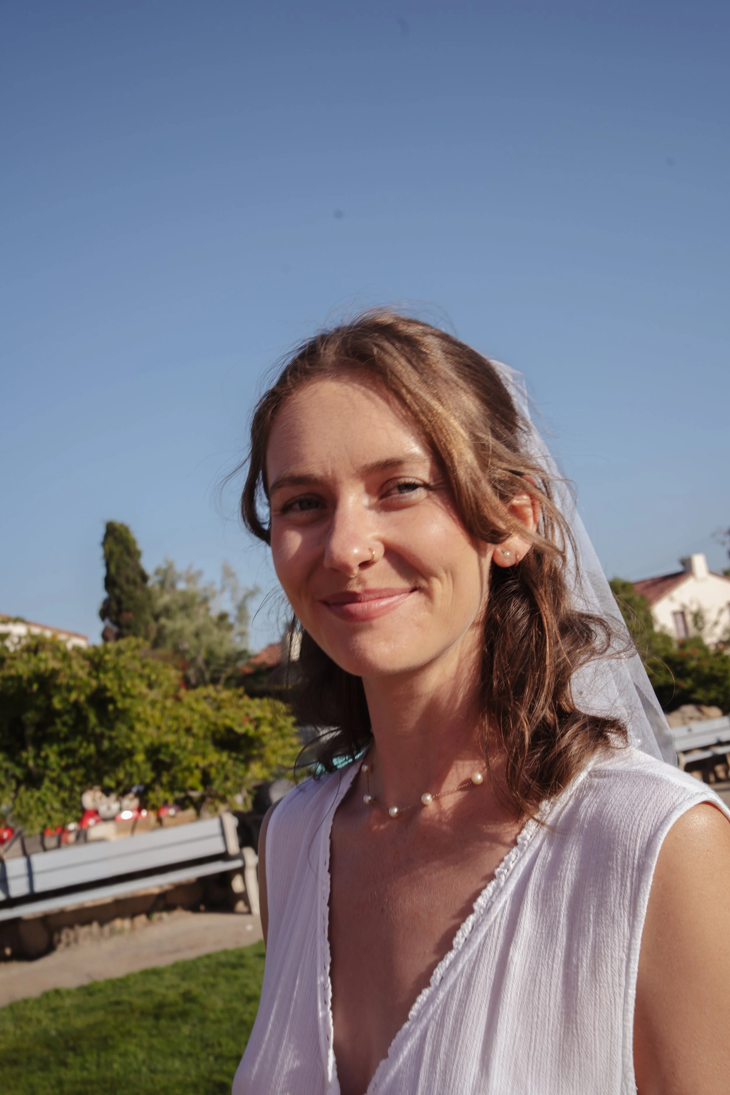 A smiling woman with brown, wavy hair and a pearl necklace standing outdoors on a sunny day, with trees, houses, and a park bench in the background.