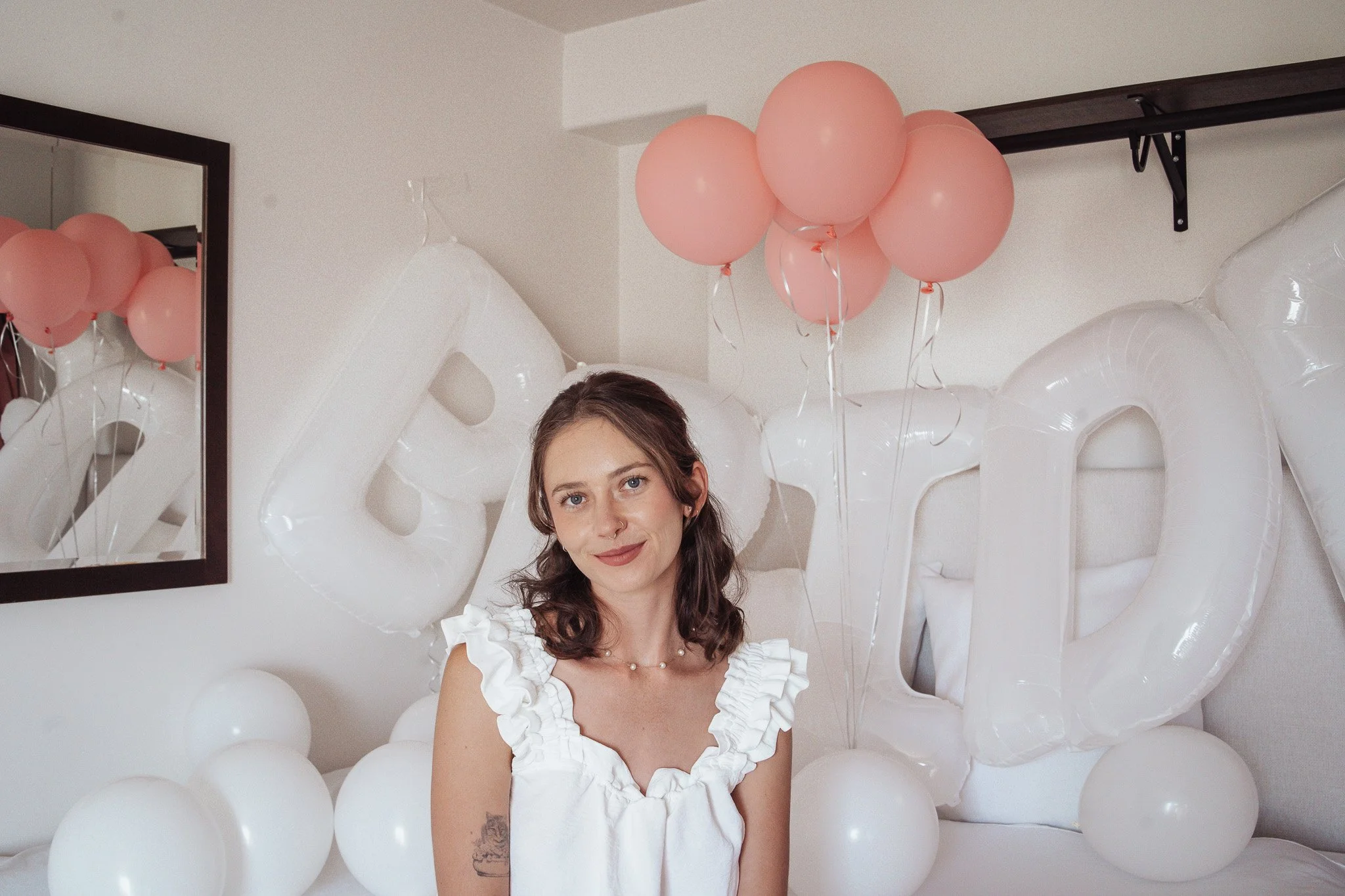 A woman with shoulder-length brown hair and light makeup is sitting on a white bed, smiling at the camera. She is wearing a white dress with ruffled sleeves and a necklace. Behind her, there are balloons in shades of pink and white, some attached to 