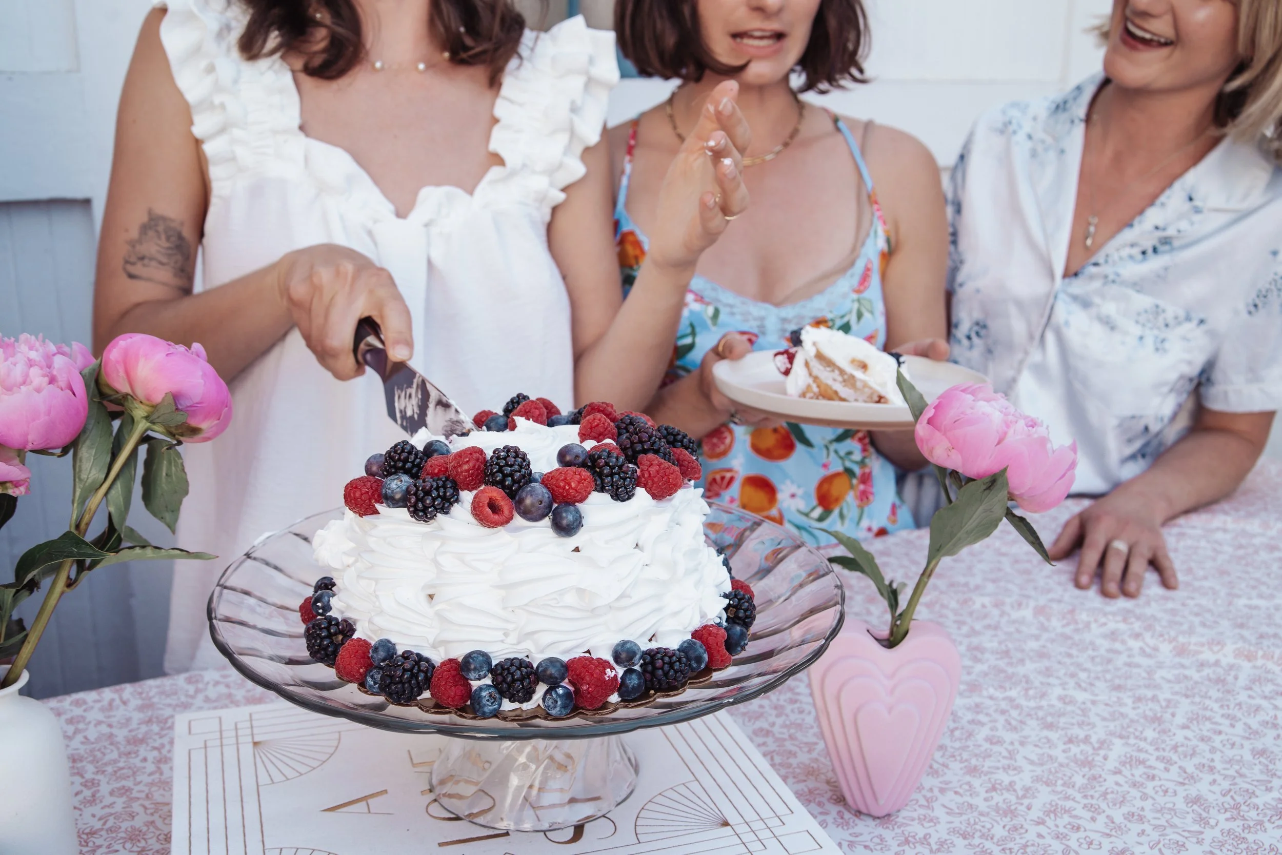 Close-up of a tiered cake topped with fresh mixed berries on a glass stand, with pink flowers and women celebrating in the background.