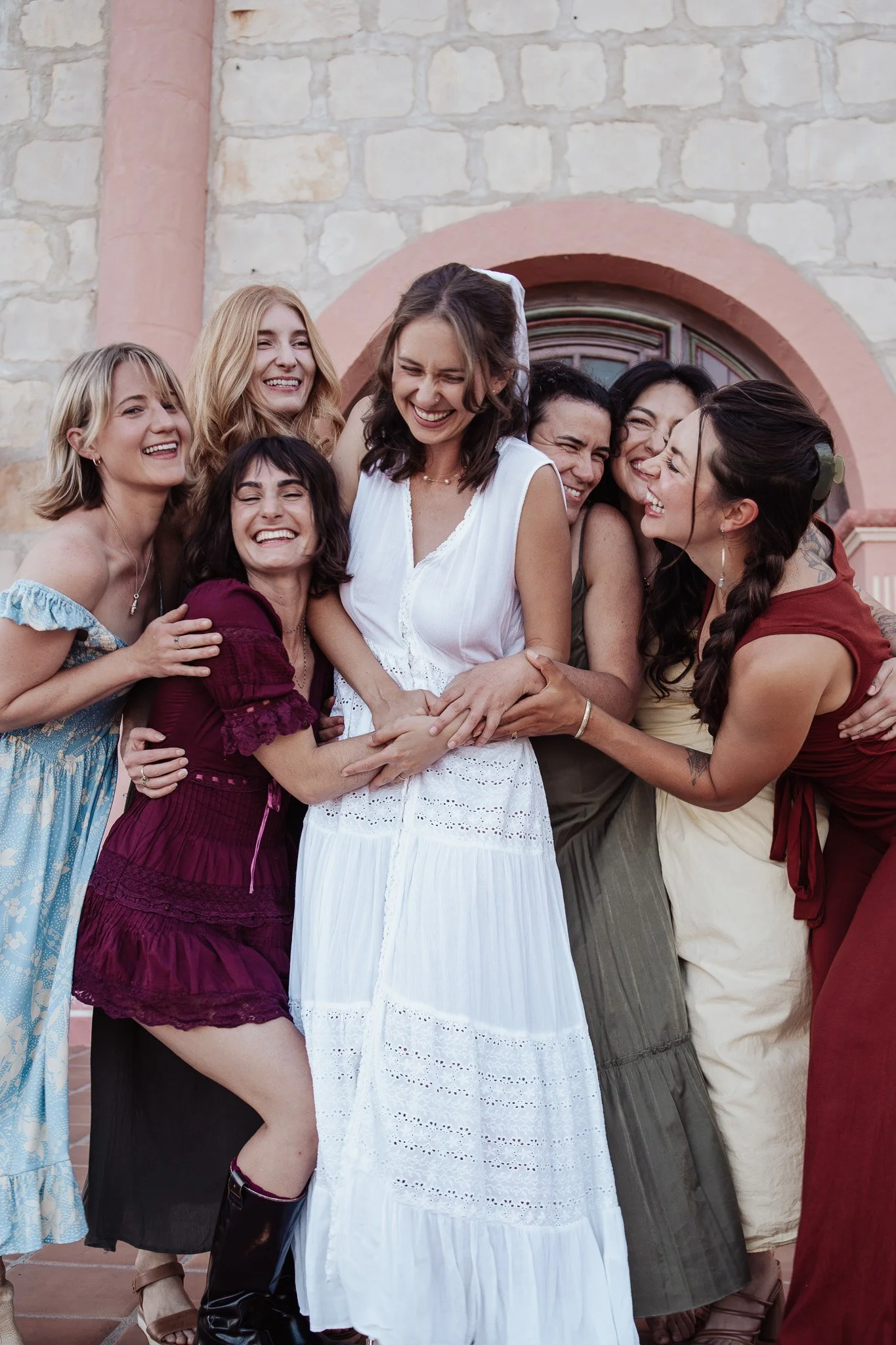 Group of women in colorful dresses hugging and laughing in front of a stone building with pink accents.