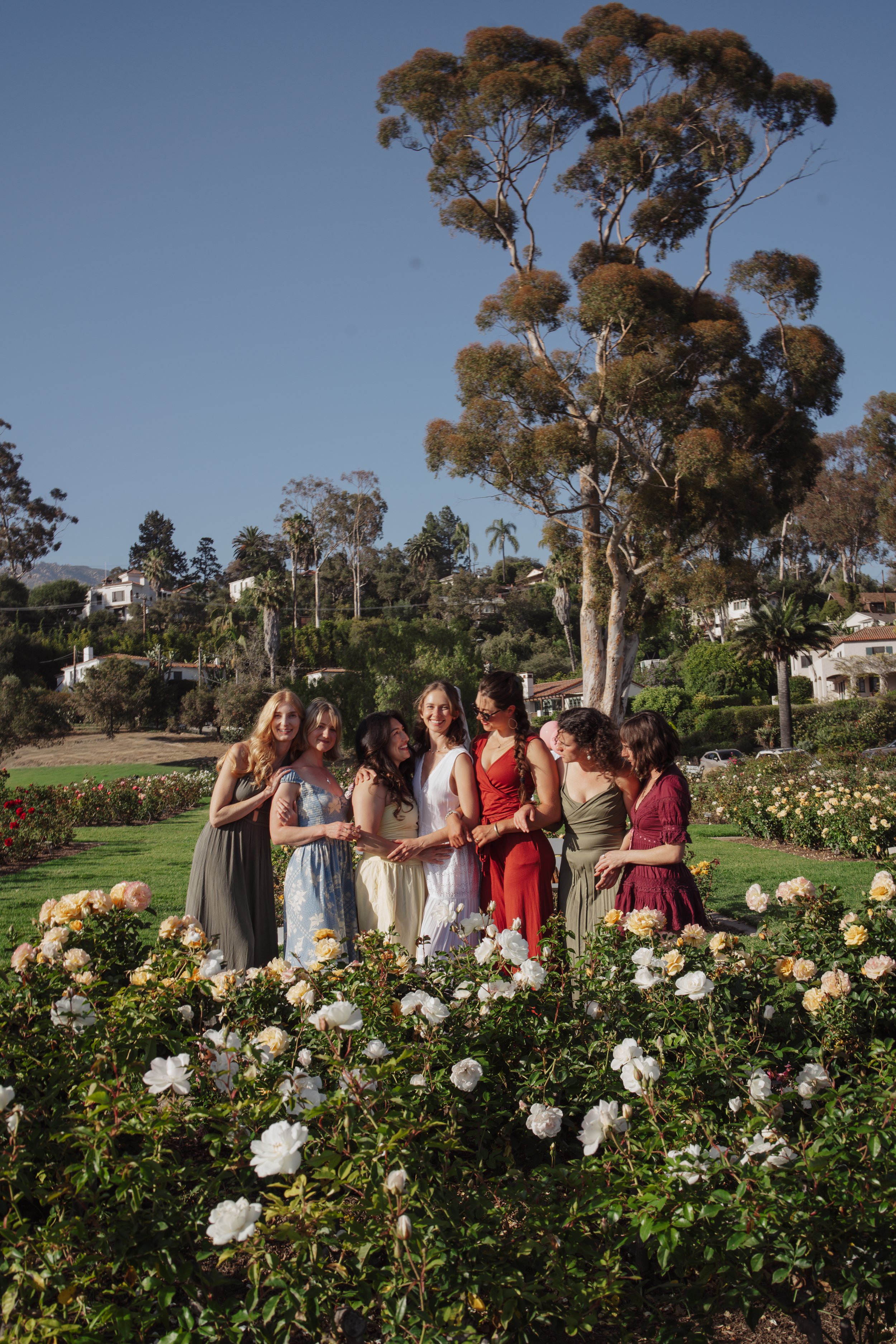 A group of women wearing colorful dresses standing outdoors at a garden wedding ceremony, with white and pink roses in the foreground and houses and trees in the background on a sunny day.