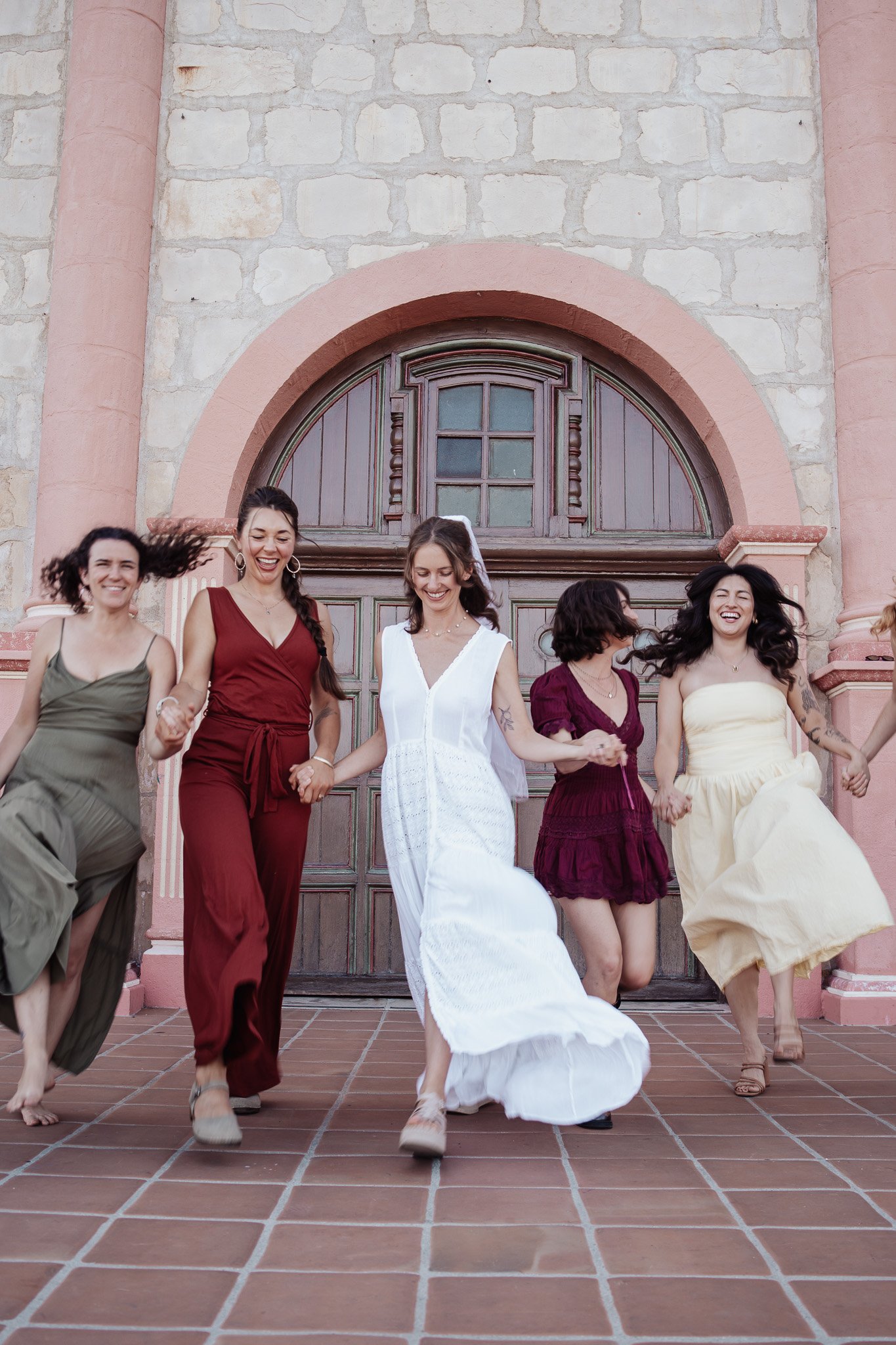 A group of women holding hands and dancing on a tiled porch in front of a stone and pink building with an arched wooden door and window.