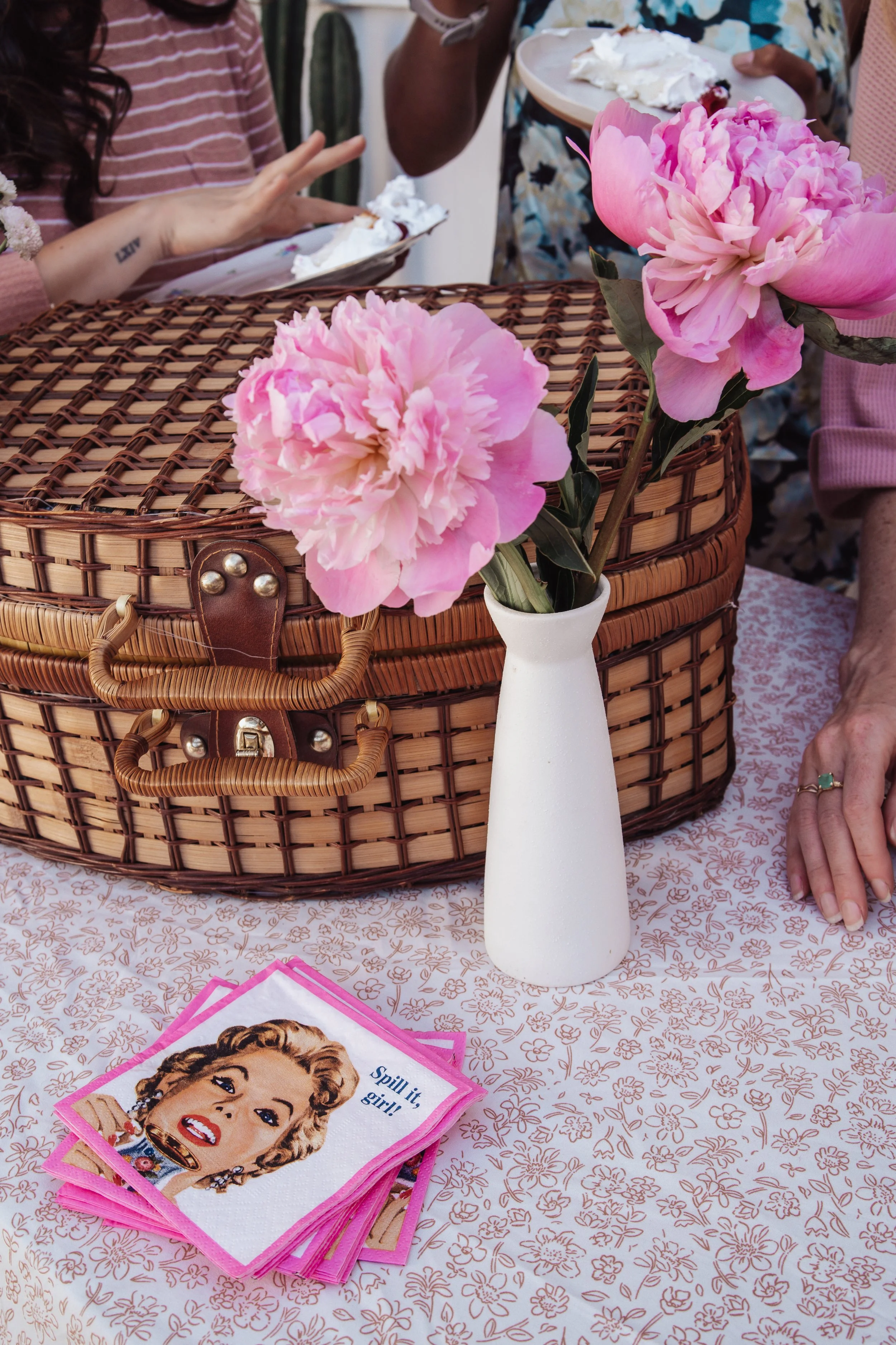 Pink peonies in a white vase placed on a table with a floral tablecloth, next to a woven basket and people enjoying dessert in the background.