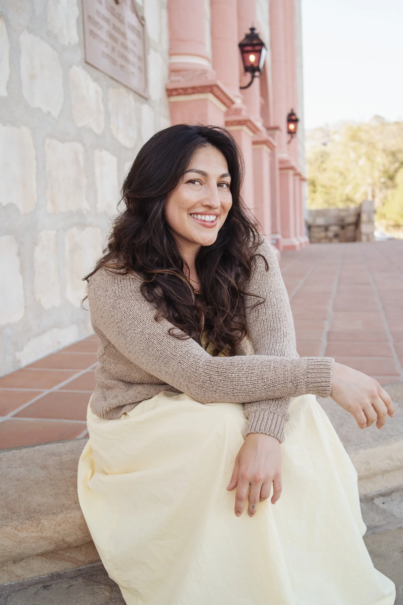 A woman with long dark hair smiling while sitting outdoors against a stone wall and pink building.