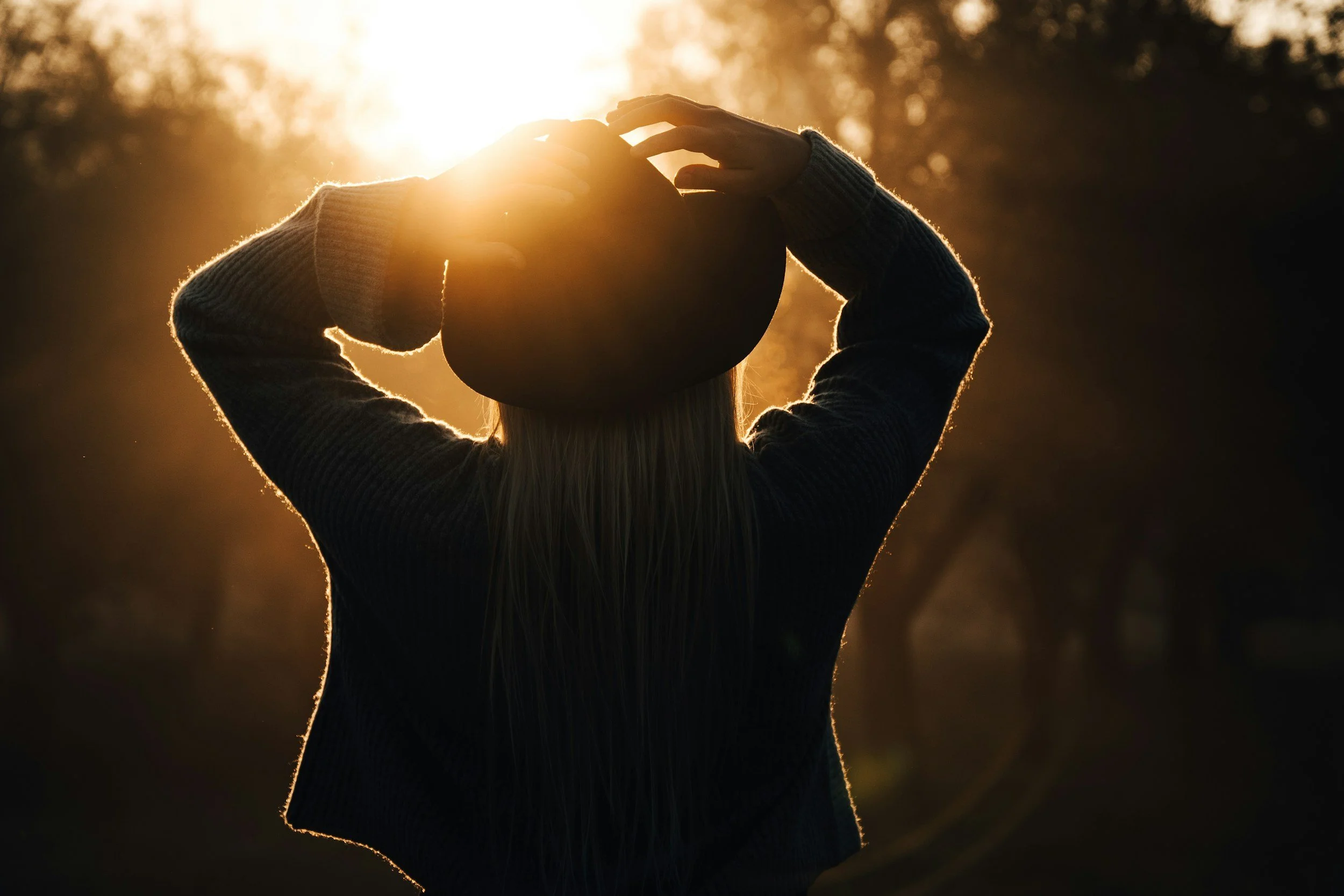 A person wearing a sweater and a hat holding a ball or helmet with both hands, standing outdoors at sunset or sunrise, with a bright light behind them.