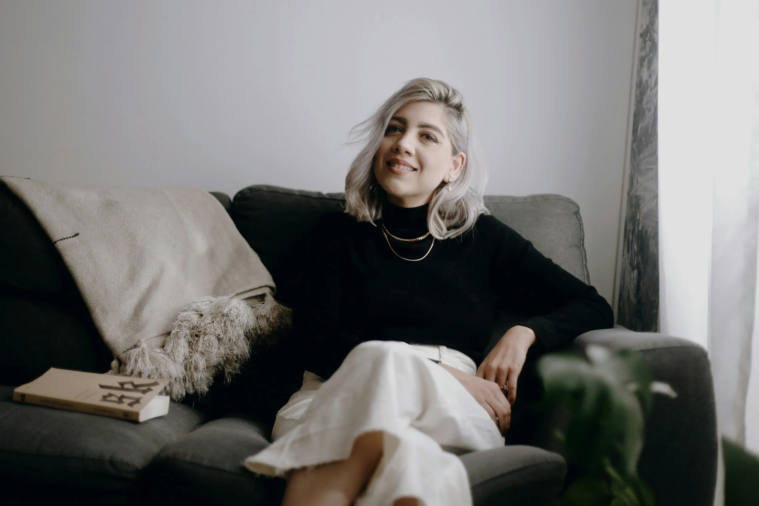A woman with blond hair and a black top sitting on a dark gray sofa near a window, smiling at the camera. There is a beige throw blanket, a plush pillow, a book, and some greenery in the foreground.