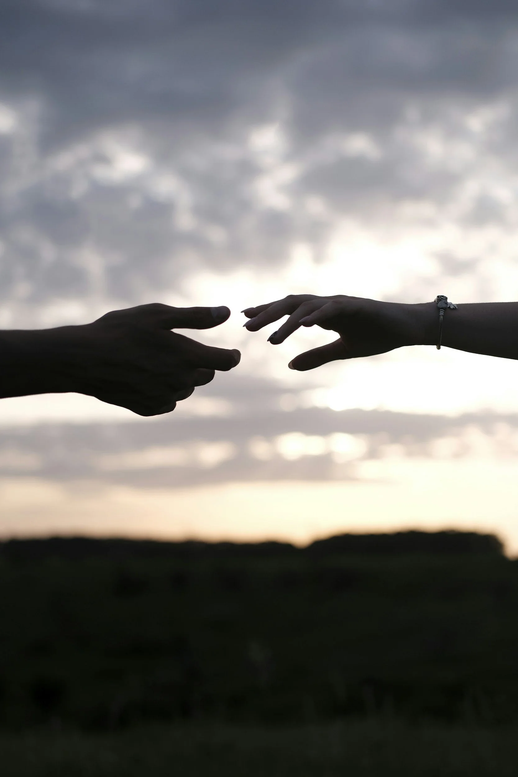 Silhouetted hands reaching towards each other against a cloudy sky at sunset, with a dark horizon line.
