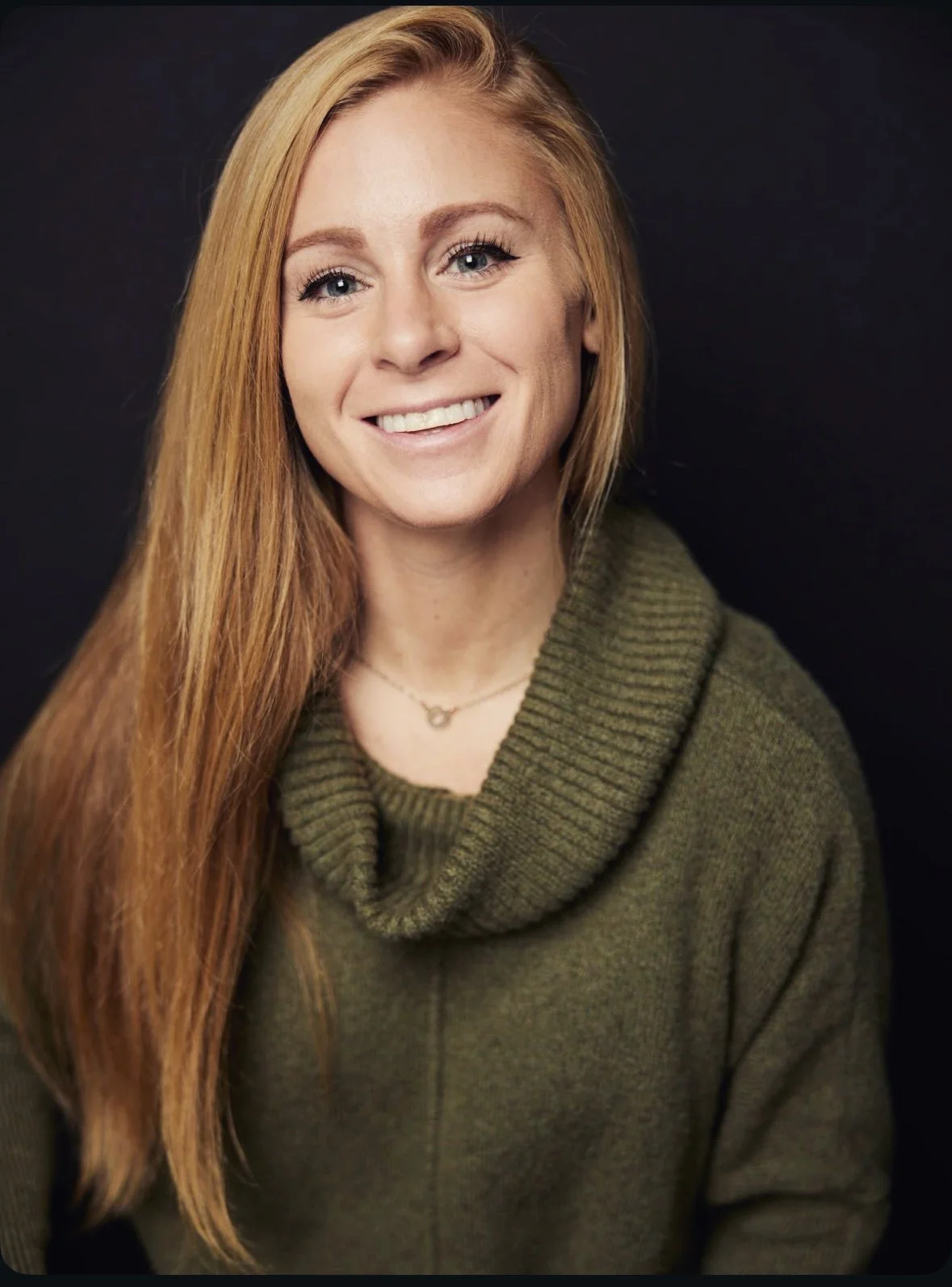 A woman with long red hair wearing an olive green knit sweater and a silver necklace, smiling against a dark background.