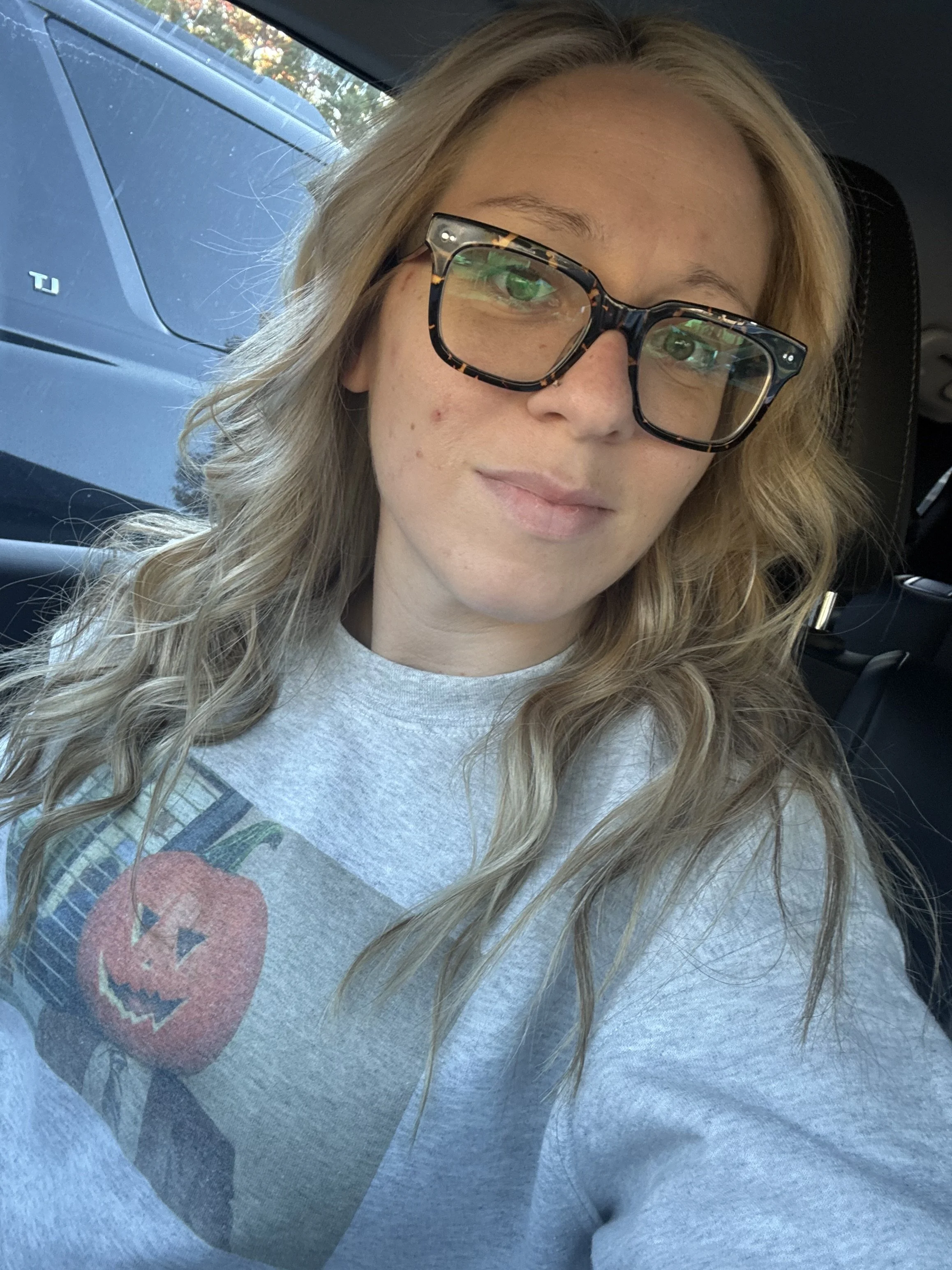A woman with long wavy blonde hair wearing dark tortoise-shell glasses and a light gray T-shirt with a graphic design, sitting inside a car.