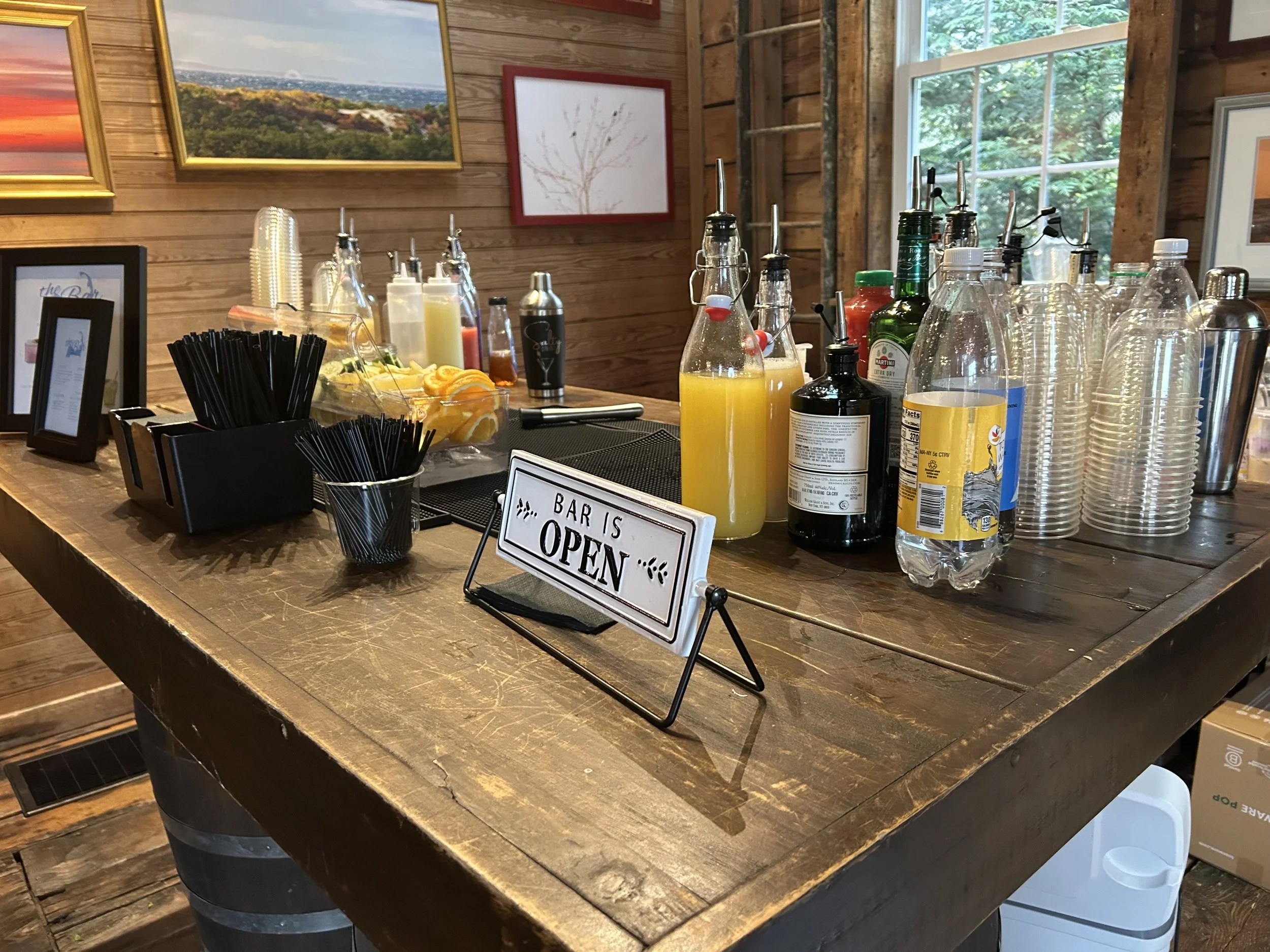 A rustic wooden bar counter with various bottles, juice, and bar tools, and a sign that reads 'Bar is Open.' There are framed landscape pictures hanging on a wooden wall and a large window showing trees outside.