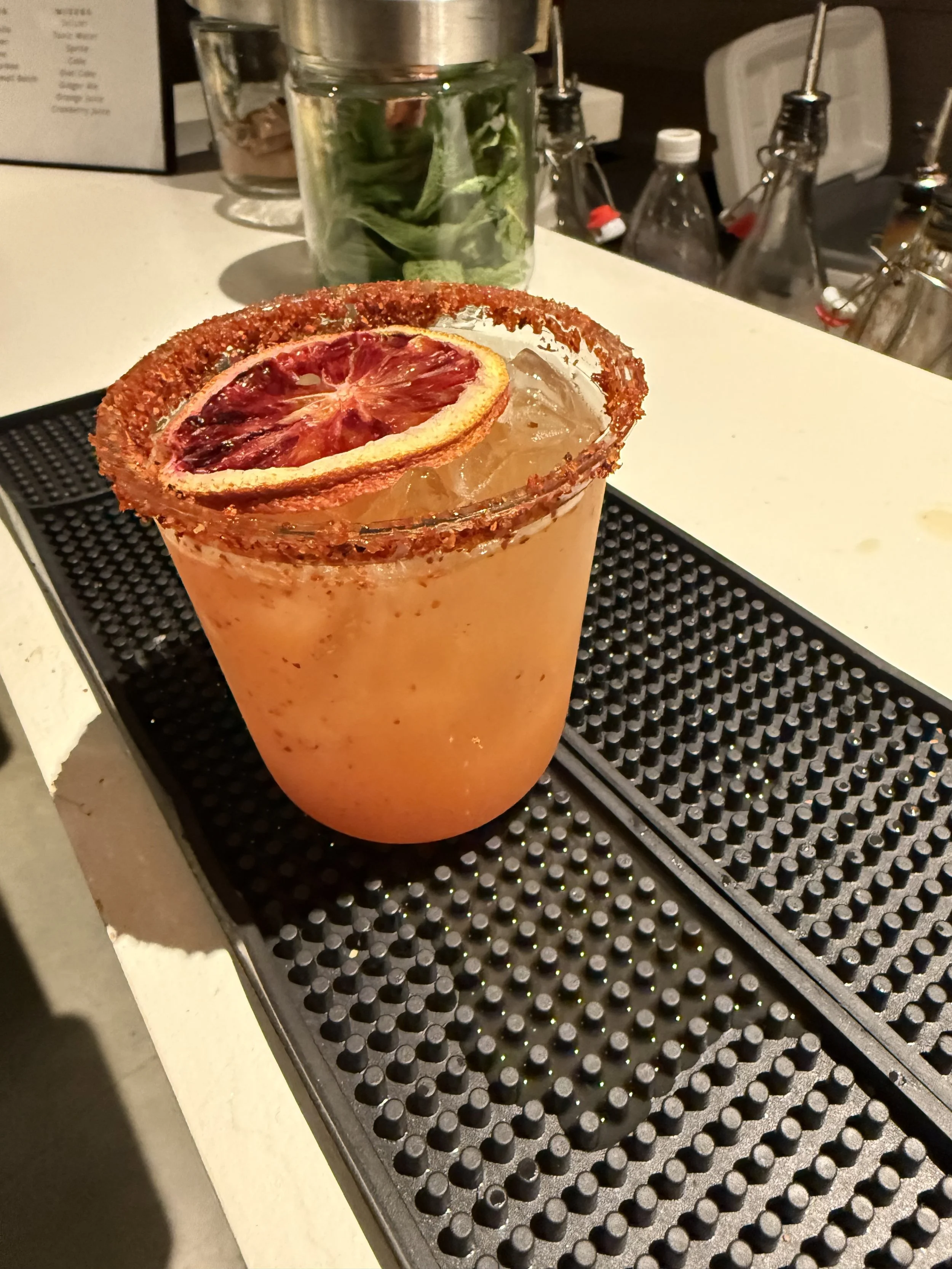 A cocktail drink garnished with a dried citrus slice, rimmed with chili powder, on a bar counter with bar tools and bottles in the background.