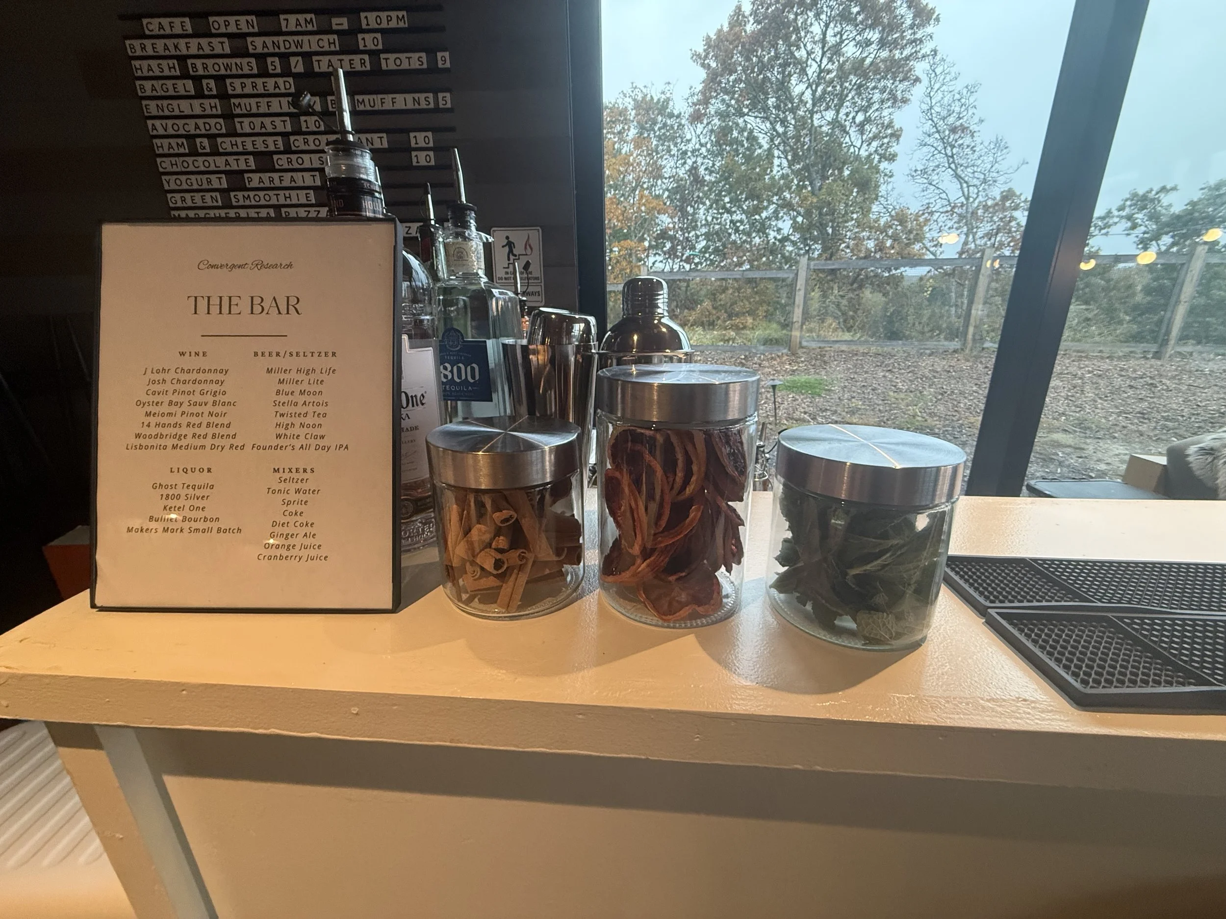 Coffee shop counter with a menu board, glass bottles of alcohol, and a glass container filled with cinnamon sticks. Large window showing trees and cloudy sky outside.
