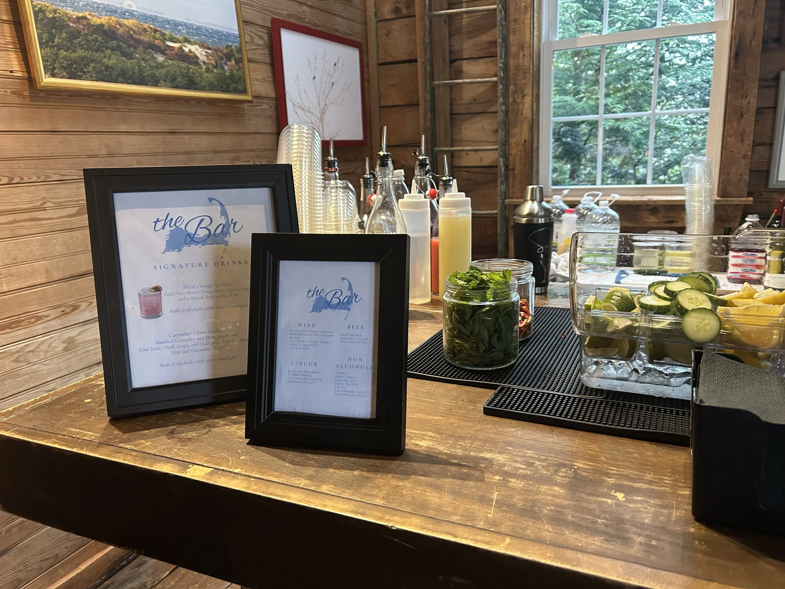 Close-up of a bar menu stand with a list of signature drinks, placed on a wooden bar counter. Behind it are bottles of syrups, a jar of mint leaves, and a glass container with sliced cucumbers and lemons, with a window showing greenery outside.